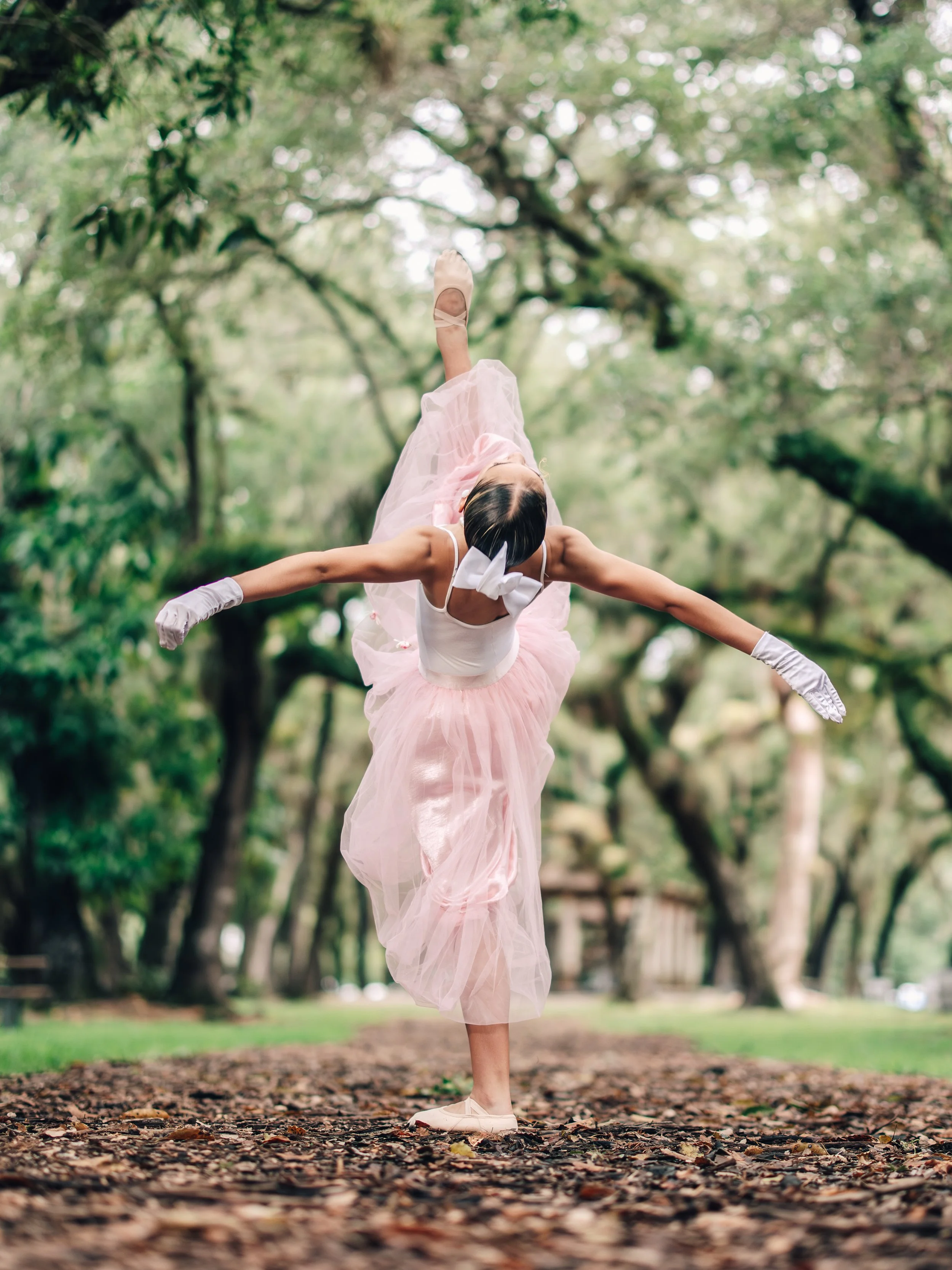 A young ballet dancer practicing outdoors on a tree-lined path, balancing on one leg with her arms extended and wearing a pink tutu and pointe shoes.