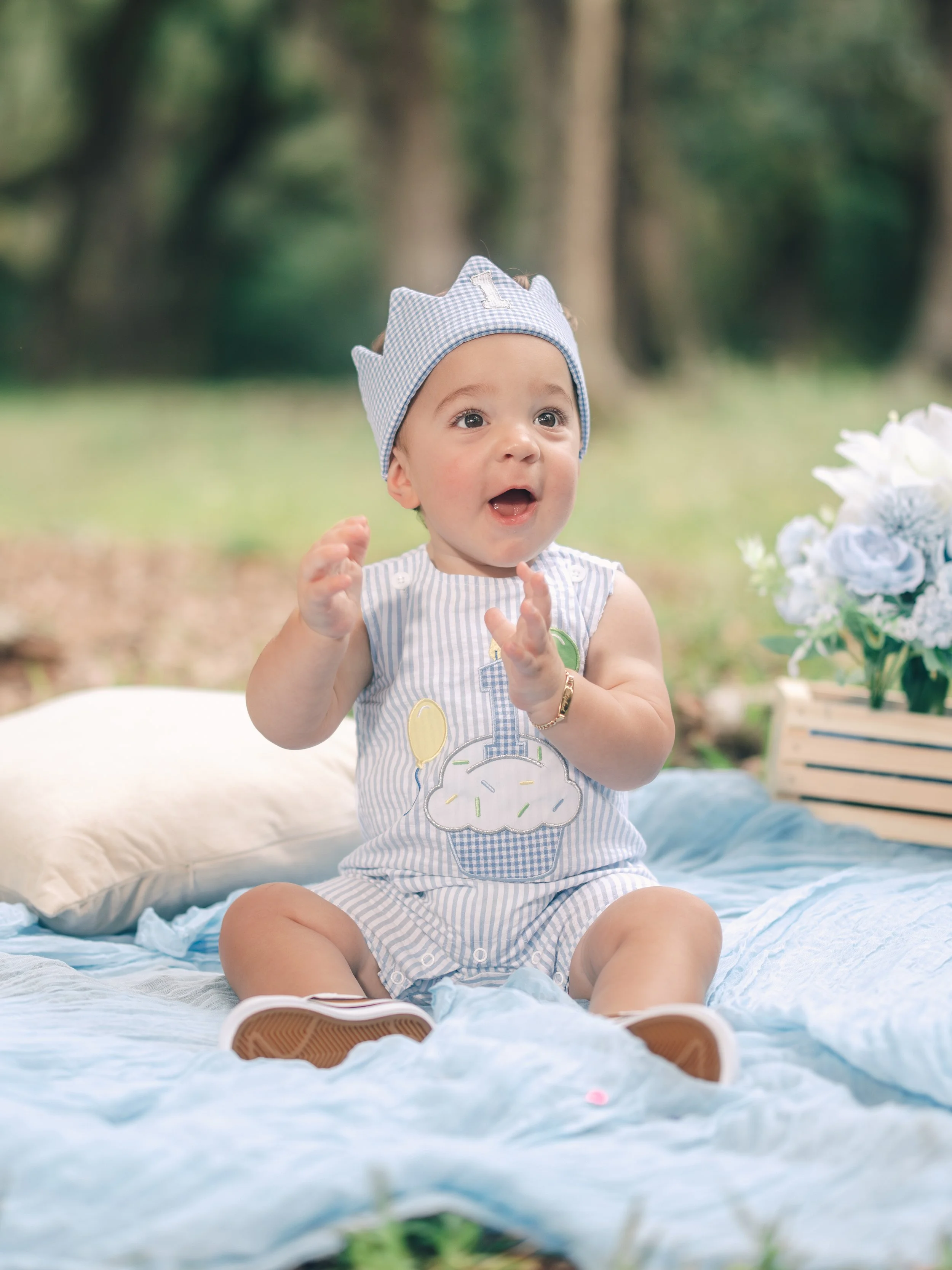 A baby sitting on a blue blanket outdoors, wearing a blue and white striped outfit with a cupcake design, a blue checkered hat with an 'I' on it, and beige shoes. There are white flowers on the right and a stack of books nearby.
