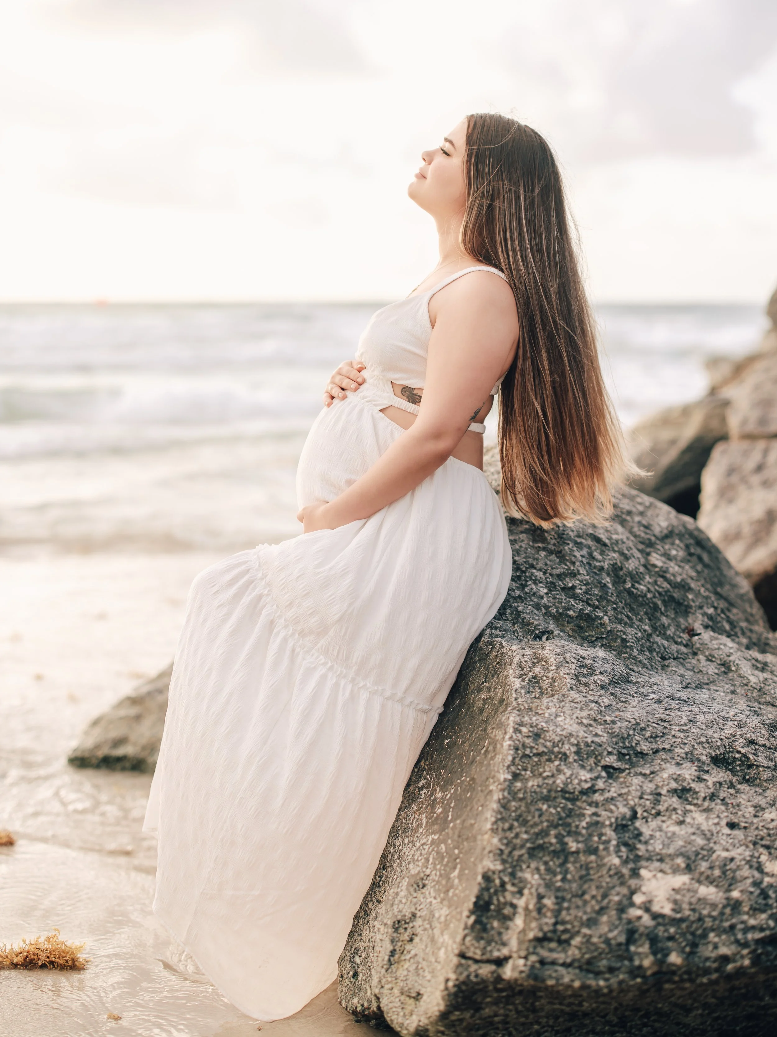 A woman in a white dress sitting on rocks by the beach with her eyes closed and smiling.