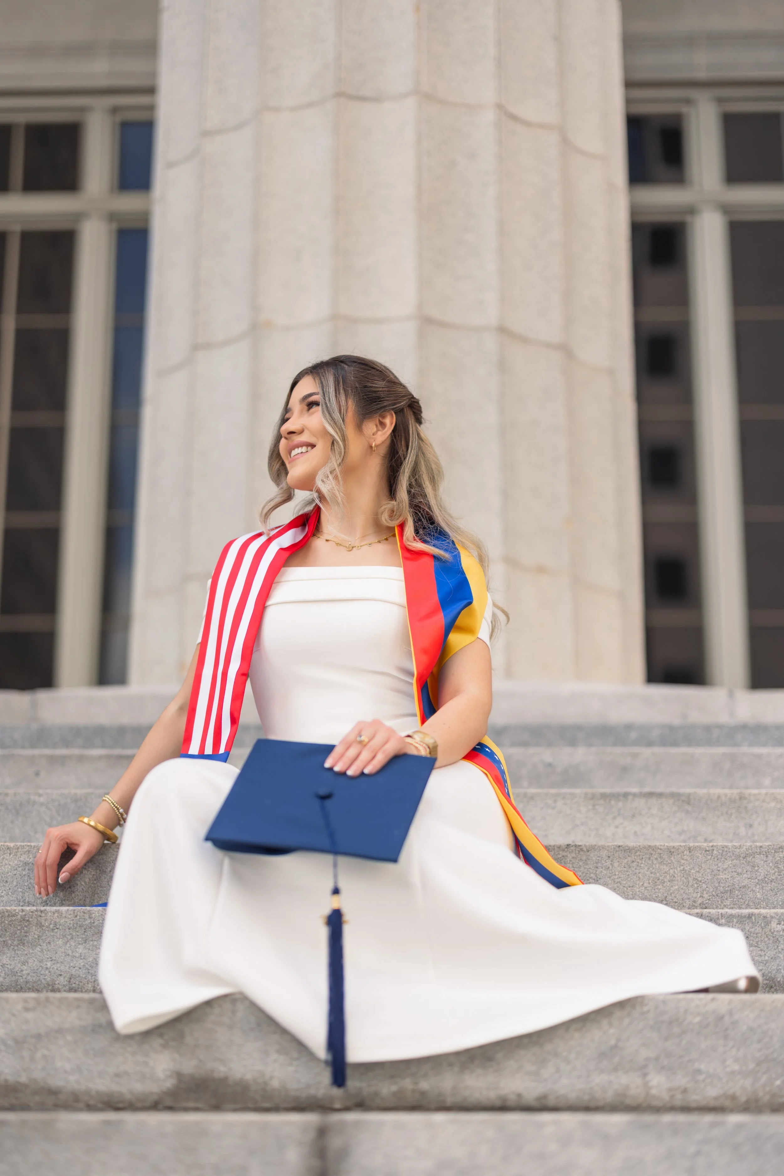 A young woman in a white dress sits on steps in front of a large building, wearing a red, yellow, and blue sash, holding a diploma, with a graduation cap on her lap, smiling and looking cheerful.