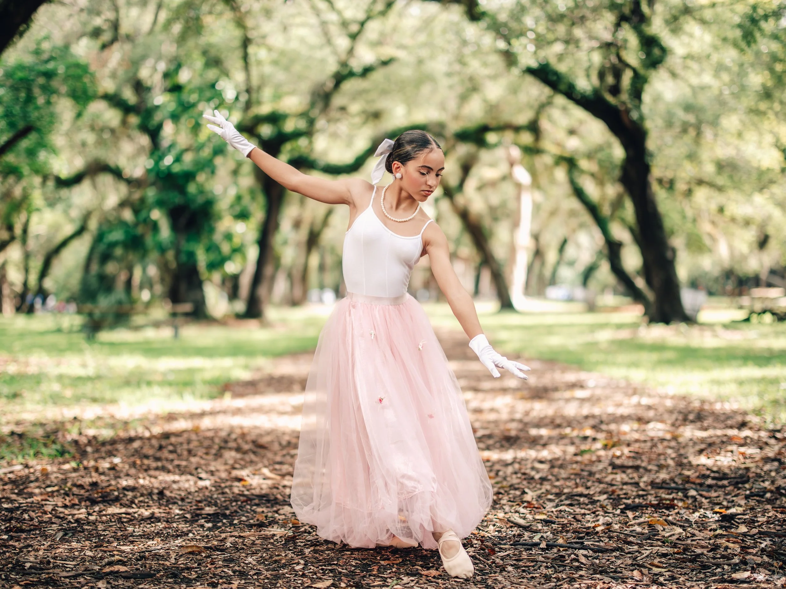A young woman dancing in a park surrounded by trees, wearing a white top, pink tulle skirt, white gloves, and pearls, with her hair tied back and a bow accessory.
