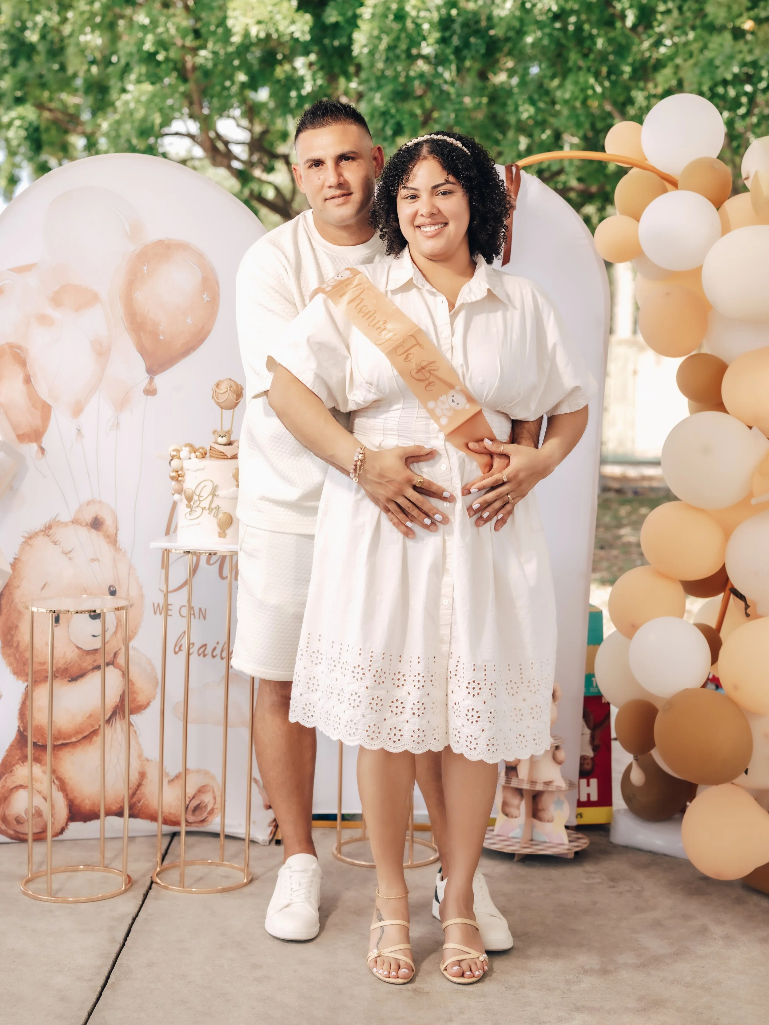 A pregnant woman and a man standing close together, smiling outdoors during a celebration with balloons and a cake decorated with teddy bears in the background. The woman wears a white dress and a sash that says 'Mommy To Be.' The man jae a white shi