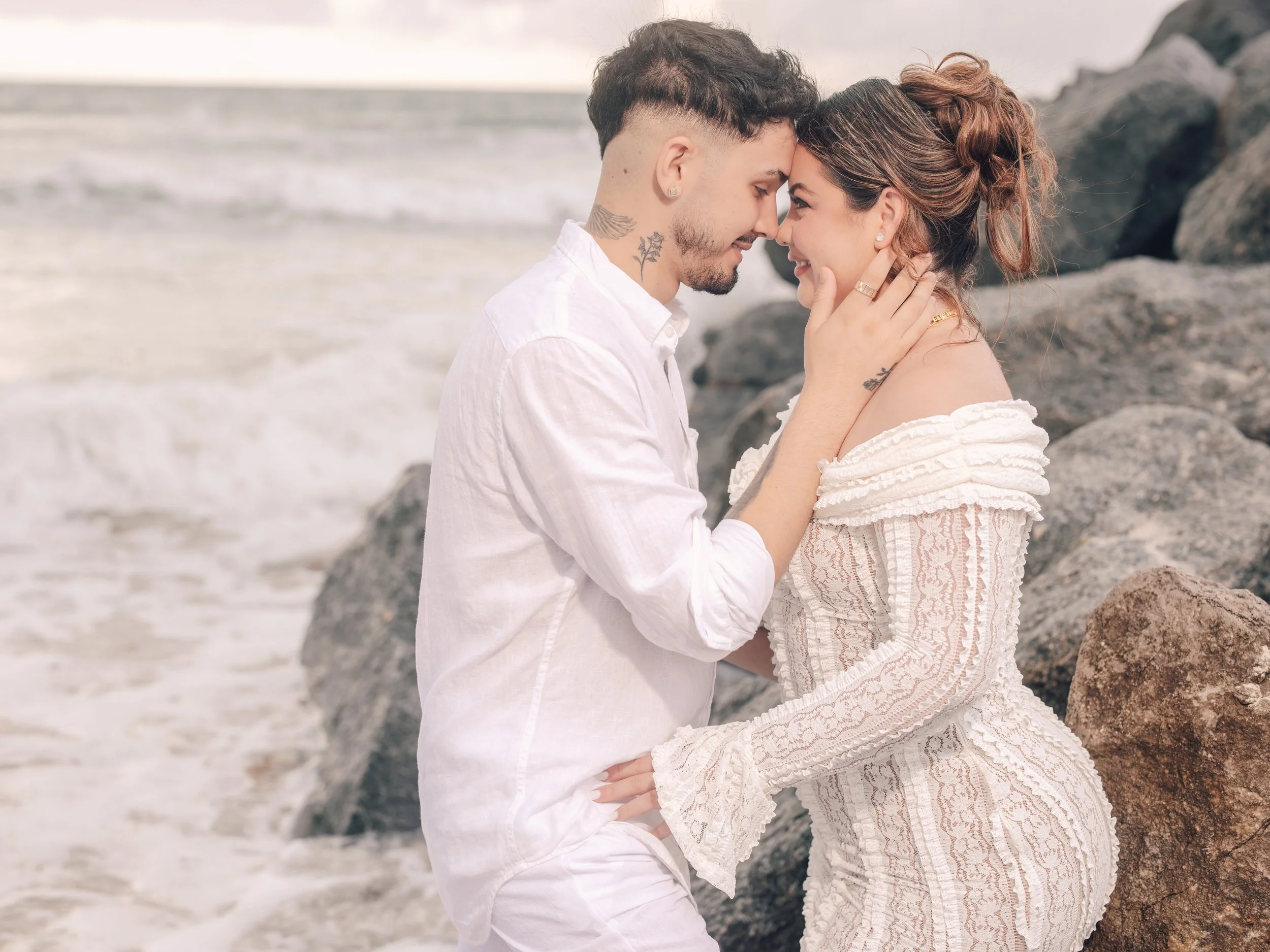 A couple is on a beach with rocks, with their foreheads touching and smiling at each other, embracing each other gently.