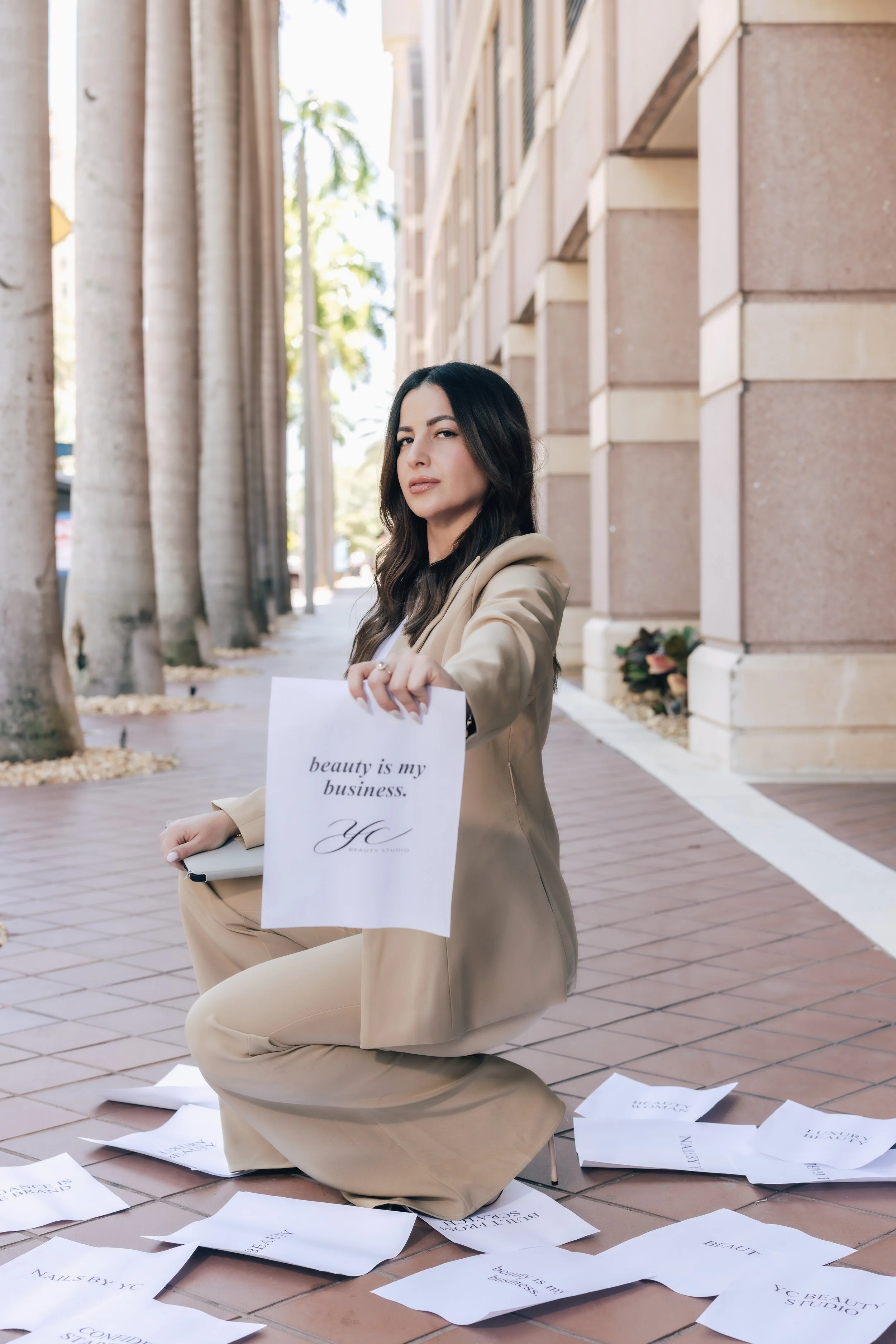 A woman dressed in beige clothing is kneeling on one knee on a tiled sidewalk outdoors, with scattered papers around her. She is holding a paper that says 'beauty is my business.' The background shows a building with large columns and palm trees.