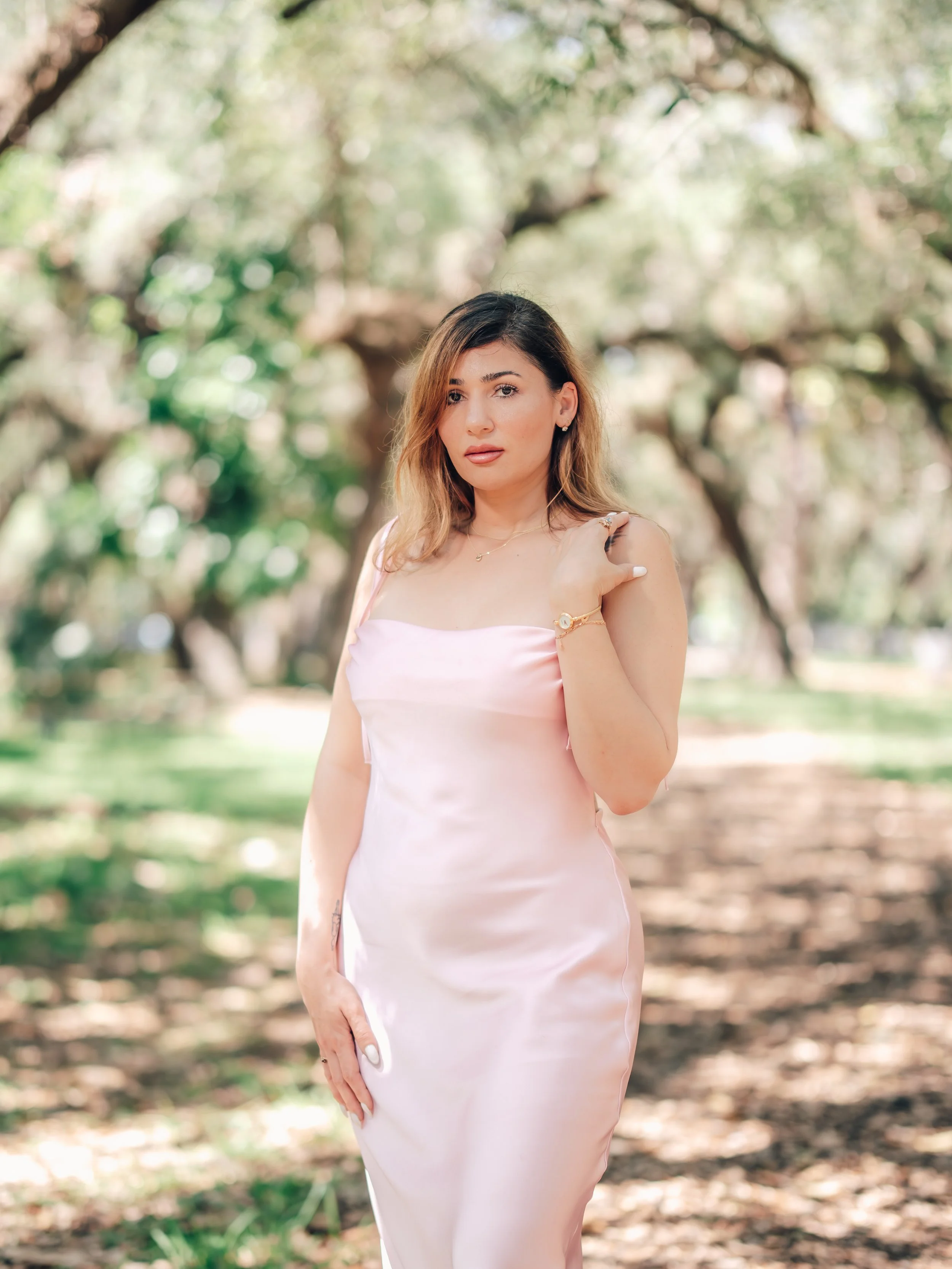 A woman with shoulder-length light brown hair wearing a pink satin dress standing outdoors among trees with blurred green foliage in the background.