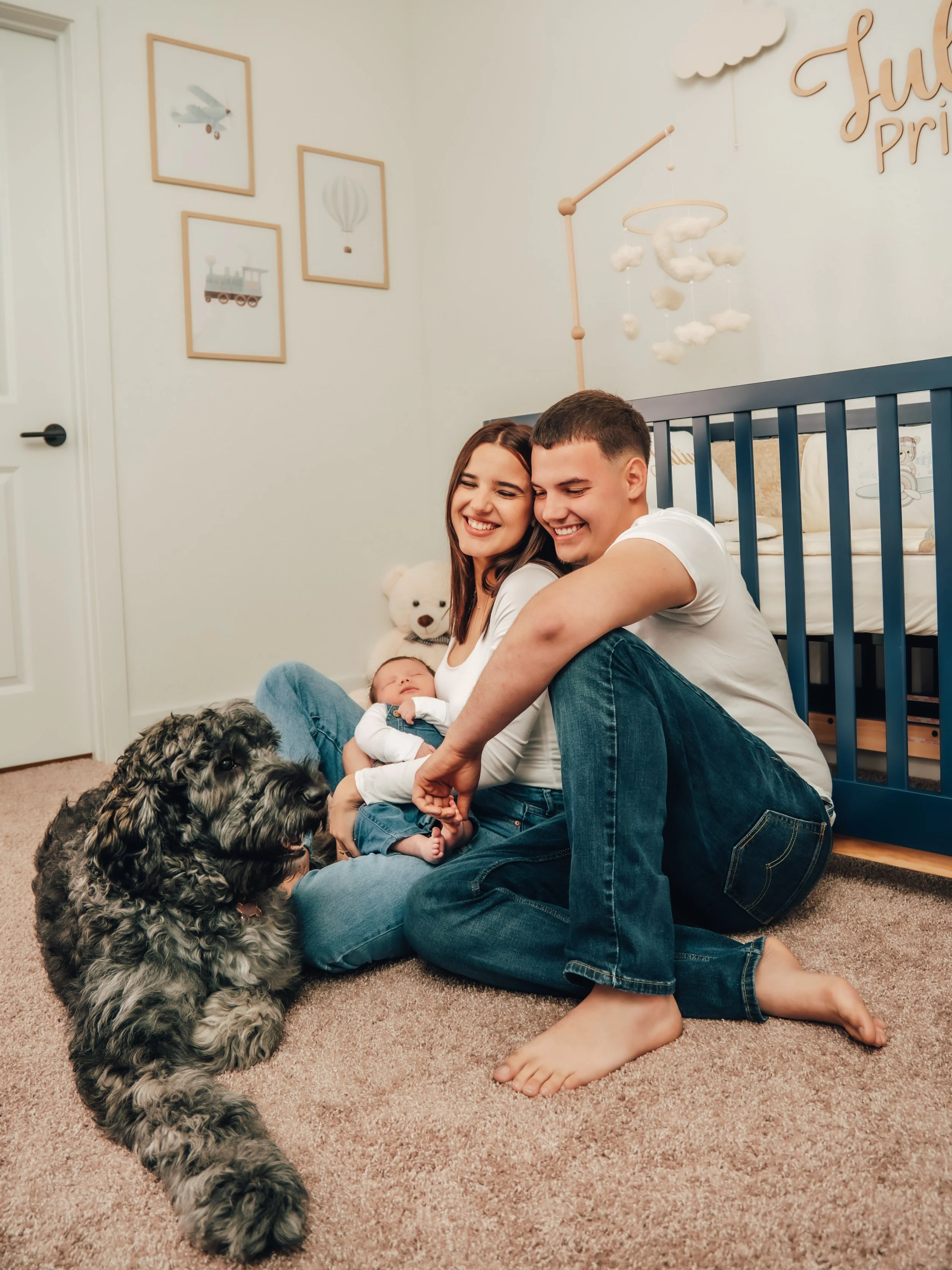 A family sitting on the carpeted floor of a nursery, smiling warmly with their newborn, a woman, a man, a puppy, and a teddy bear, with moon cloud mobile and framed pictures on the wall.
