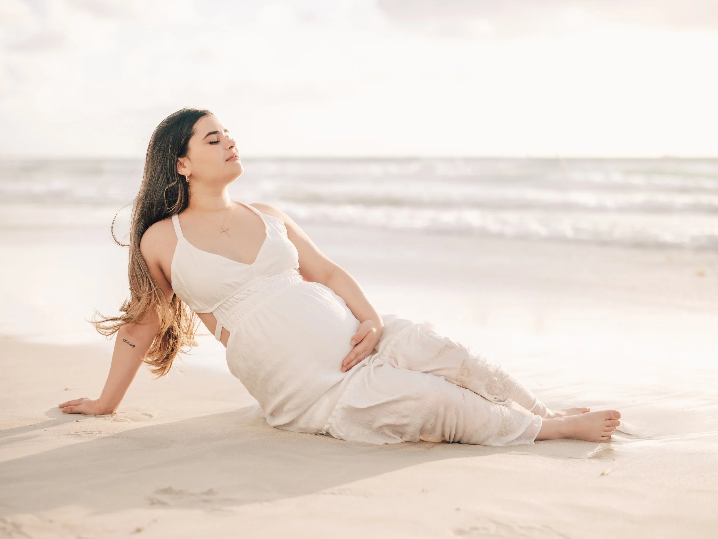 A pregnant woman in a white dress relaxing on the sandy beach with the ocean in the background, her eyes closed, enjoying the moment.