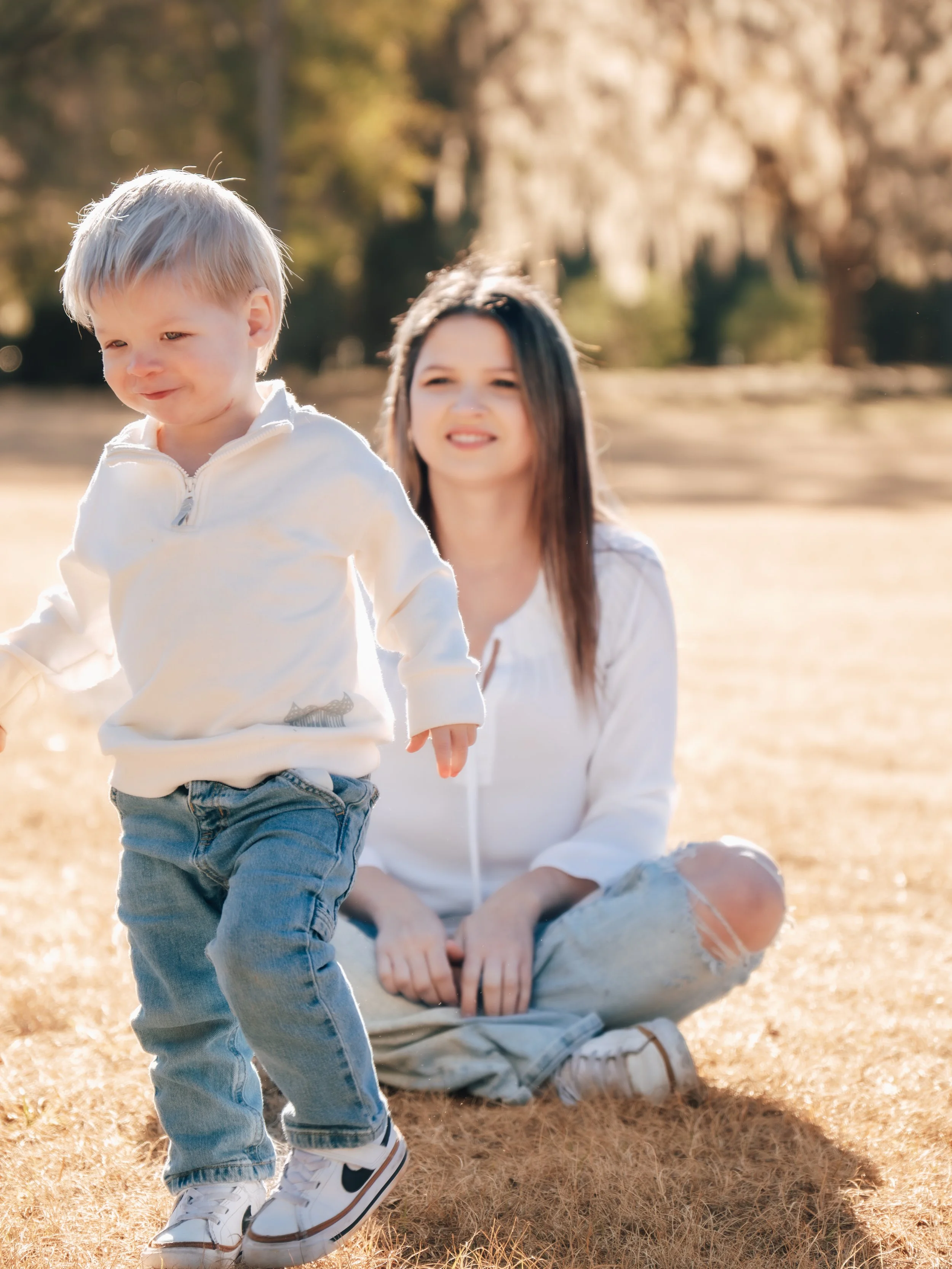 A woman sitting on the grass outdoors with a young boy in front, both wearing casual clothes on a sunny day in a park in Ocala, FL