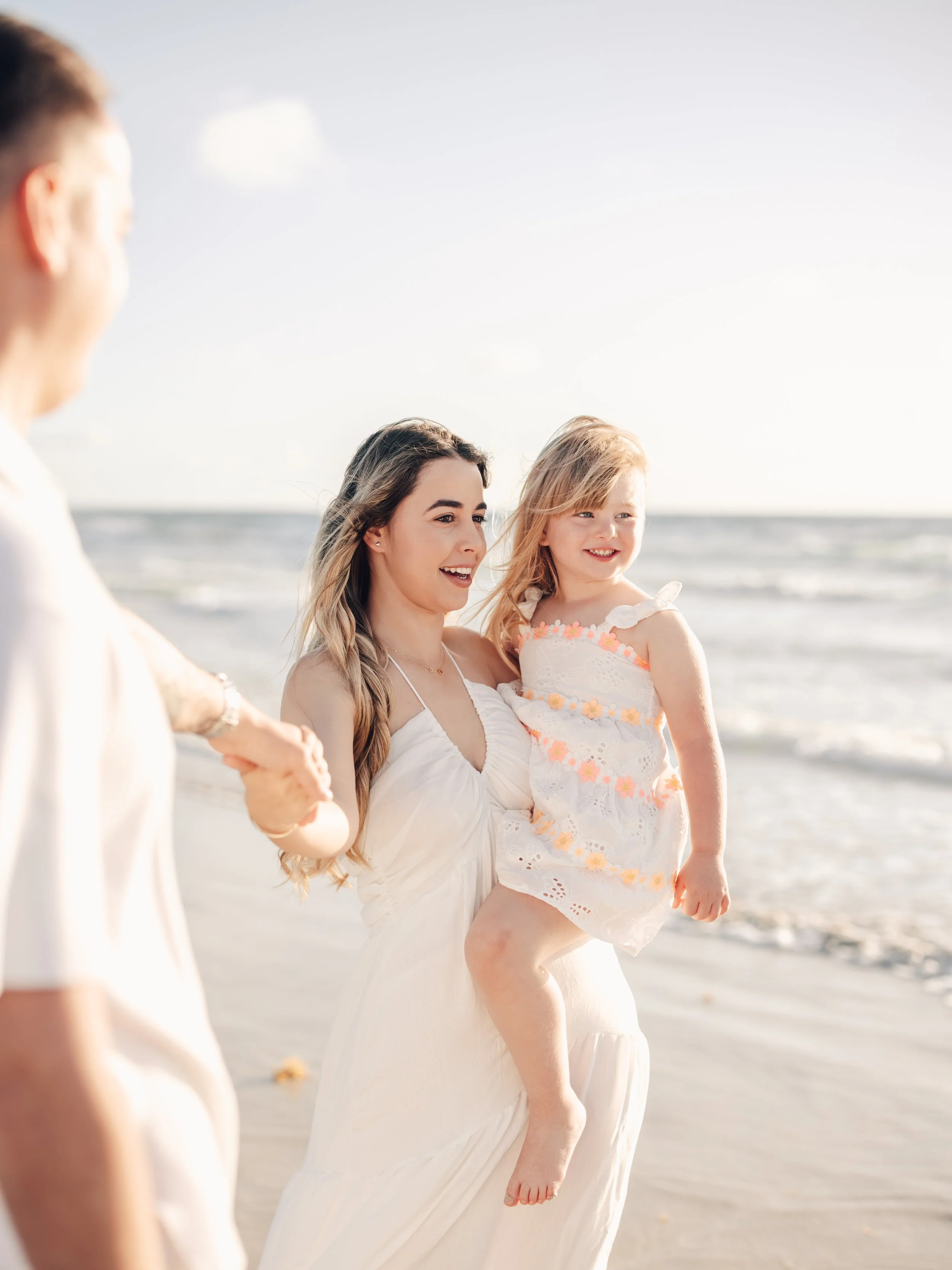 A woman in a white dress holding a young girl with blonde hair on a beach, with others holding hands in a circle. The sun is shining, and the ocean waves are visible in the background.