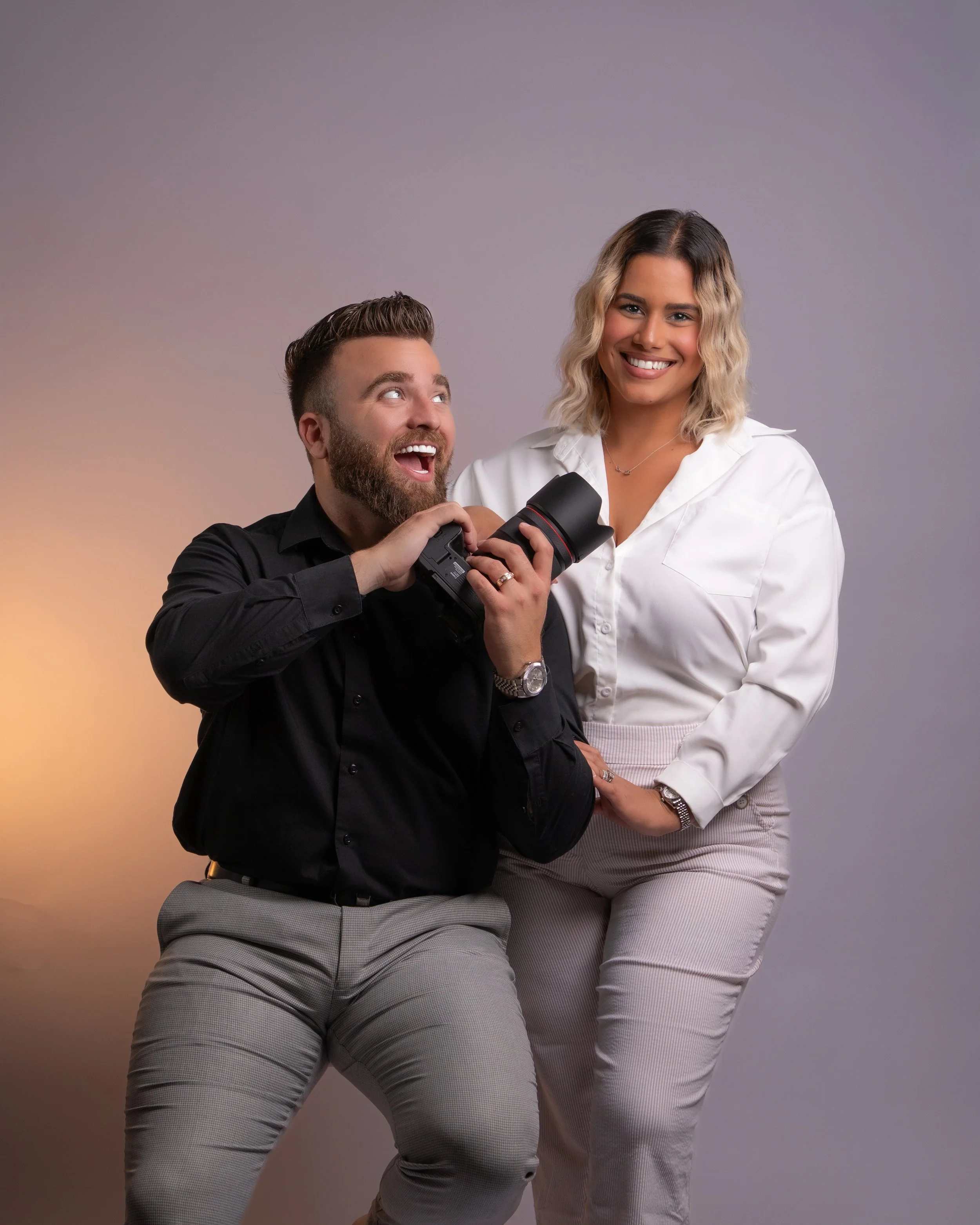A man sitting and smiling while holding a camera, with a woman standing beside him, smiling at the camera, against a plain gray background.