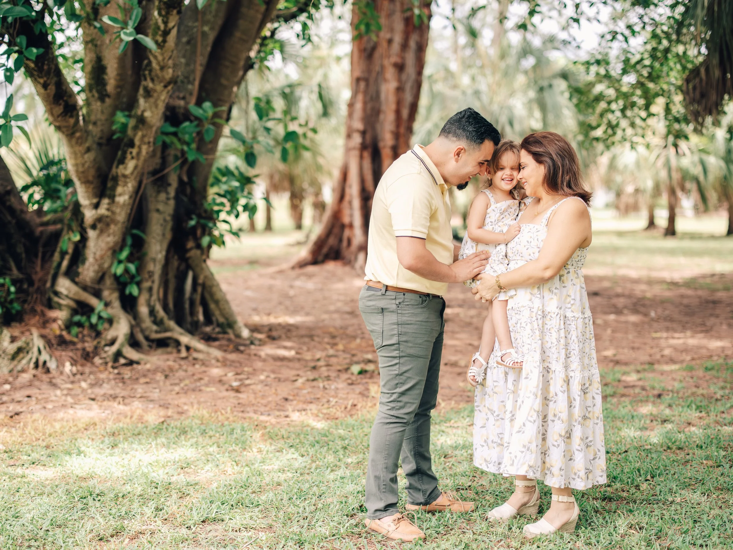 A family of three, a man, a woman, and a young girl, are outdoors in a forested area, smiling and touching heads as they share a joyful moment together.