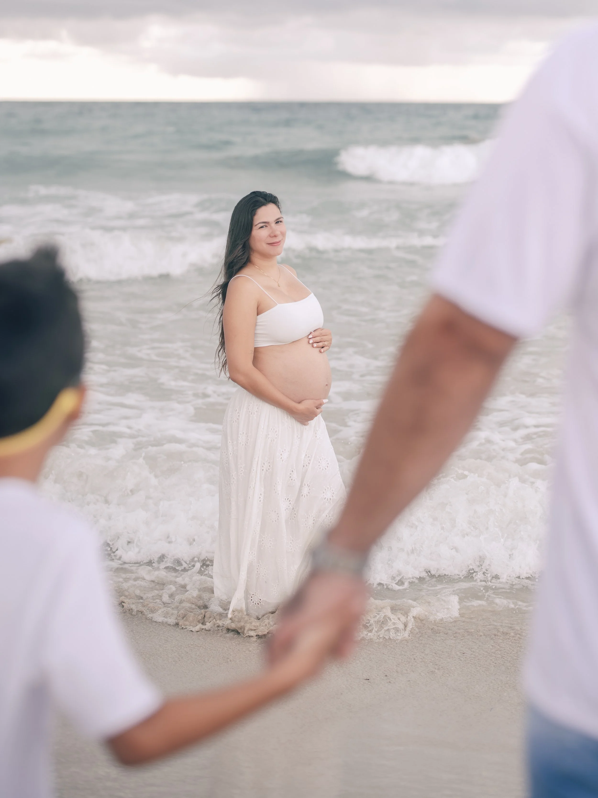 A pregnant woman standing on the beach near the water, holding her belly, with a couple holding hands in the foreground. The woman is smiling and wearing a white dress, and beach waves are in the background under a cloudy sky.
