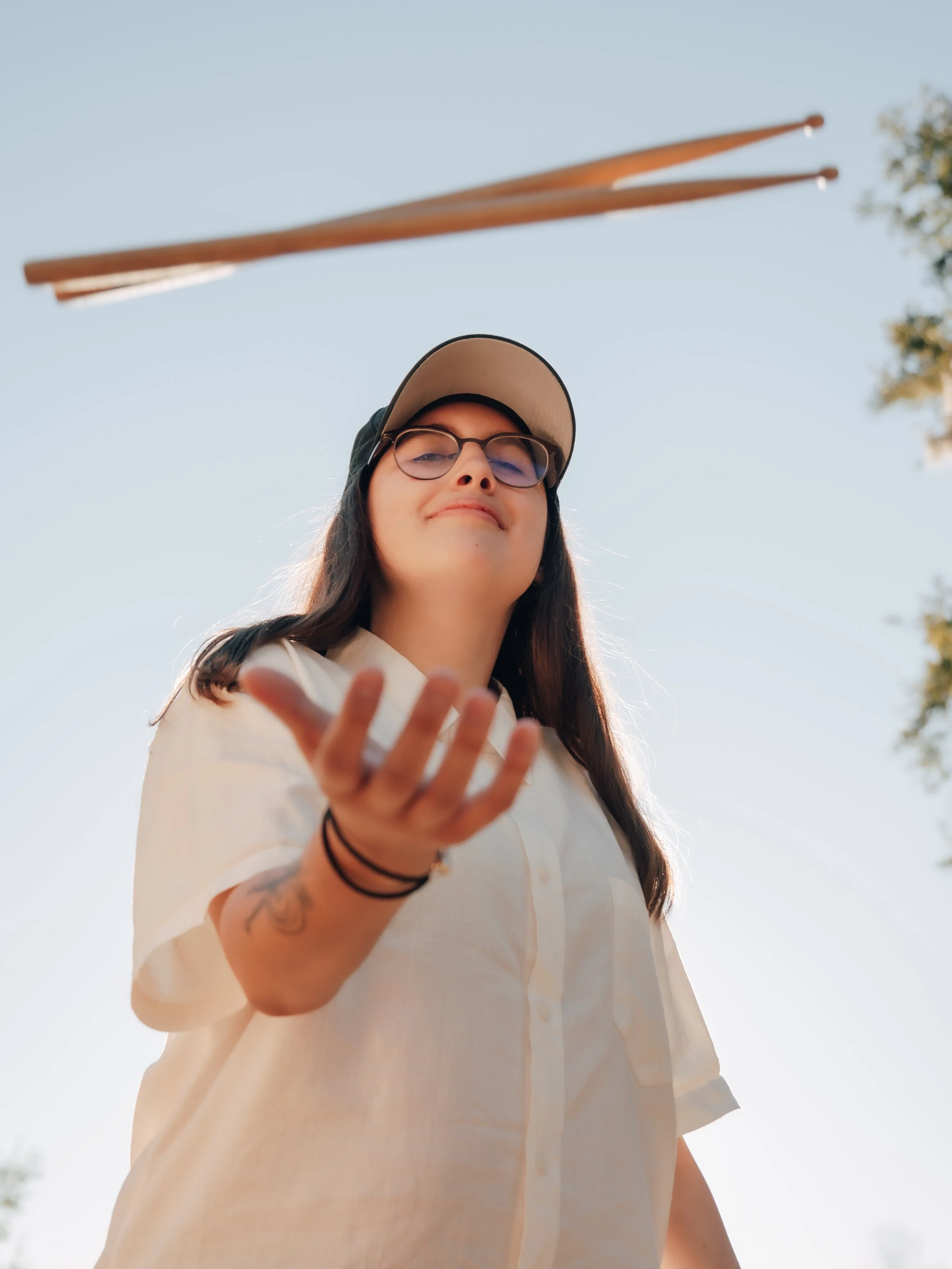 A young woman wearing glasses, a baseball cap, and a white shirt is tossing a frisbee in the air outdoors against a clear blue sky.