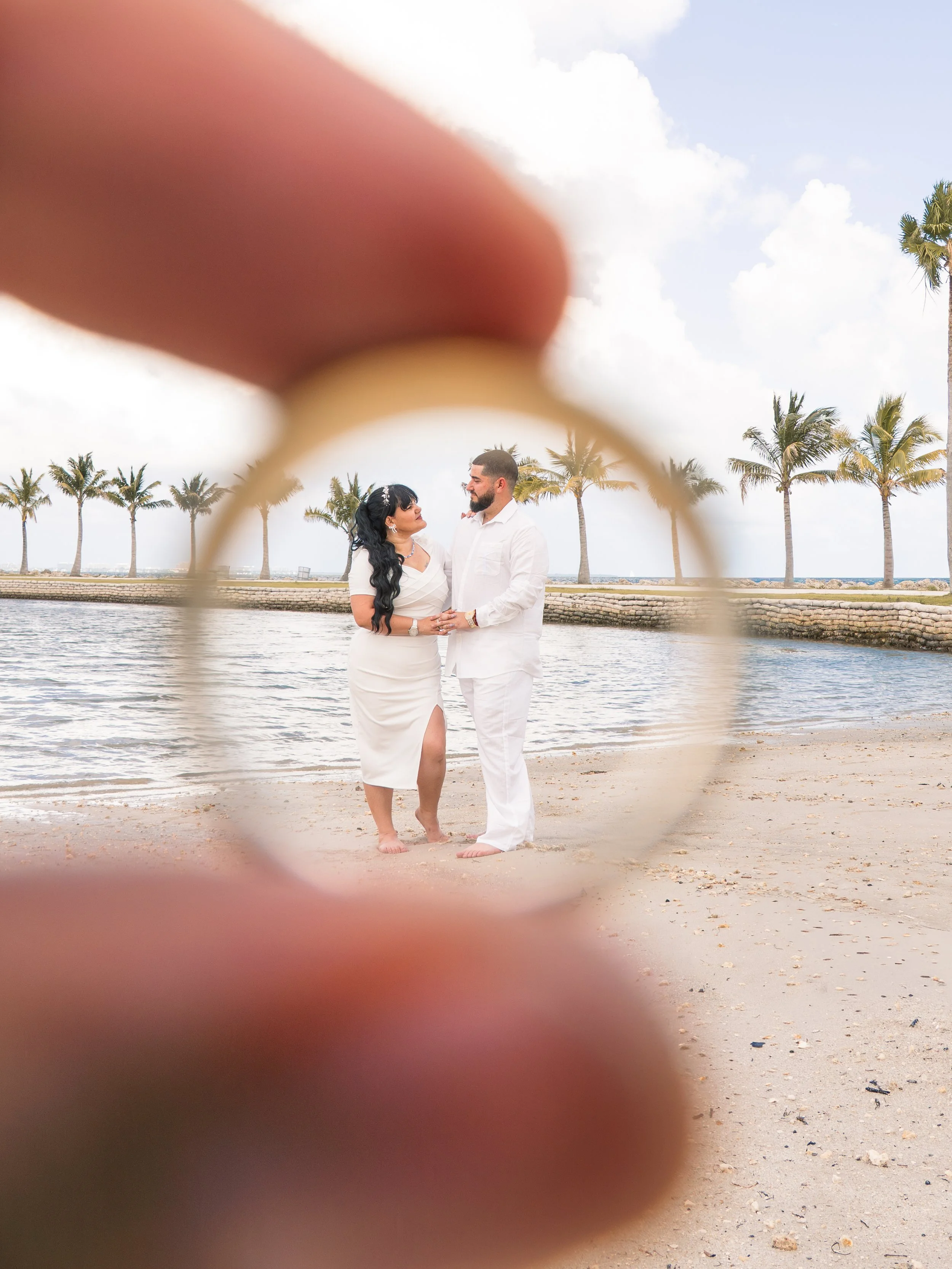 A couple stands by the water on a beach with palm trees in the background, through a blurred finger in the foreground.