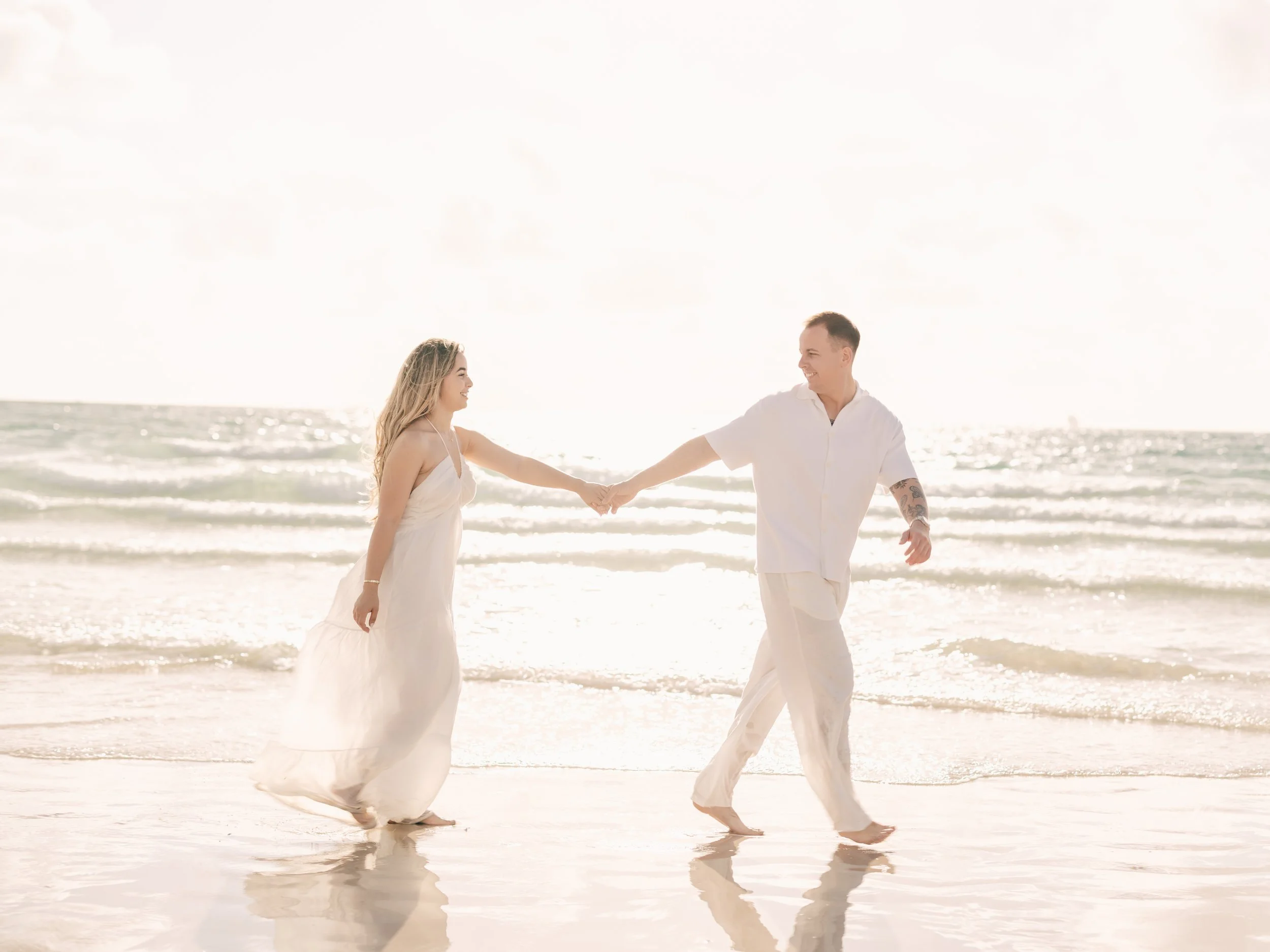 A smiling couple holding hands and walking on the beach, dressed in white, with the sun shining brightly in the sky and reflections on the water.