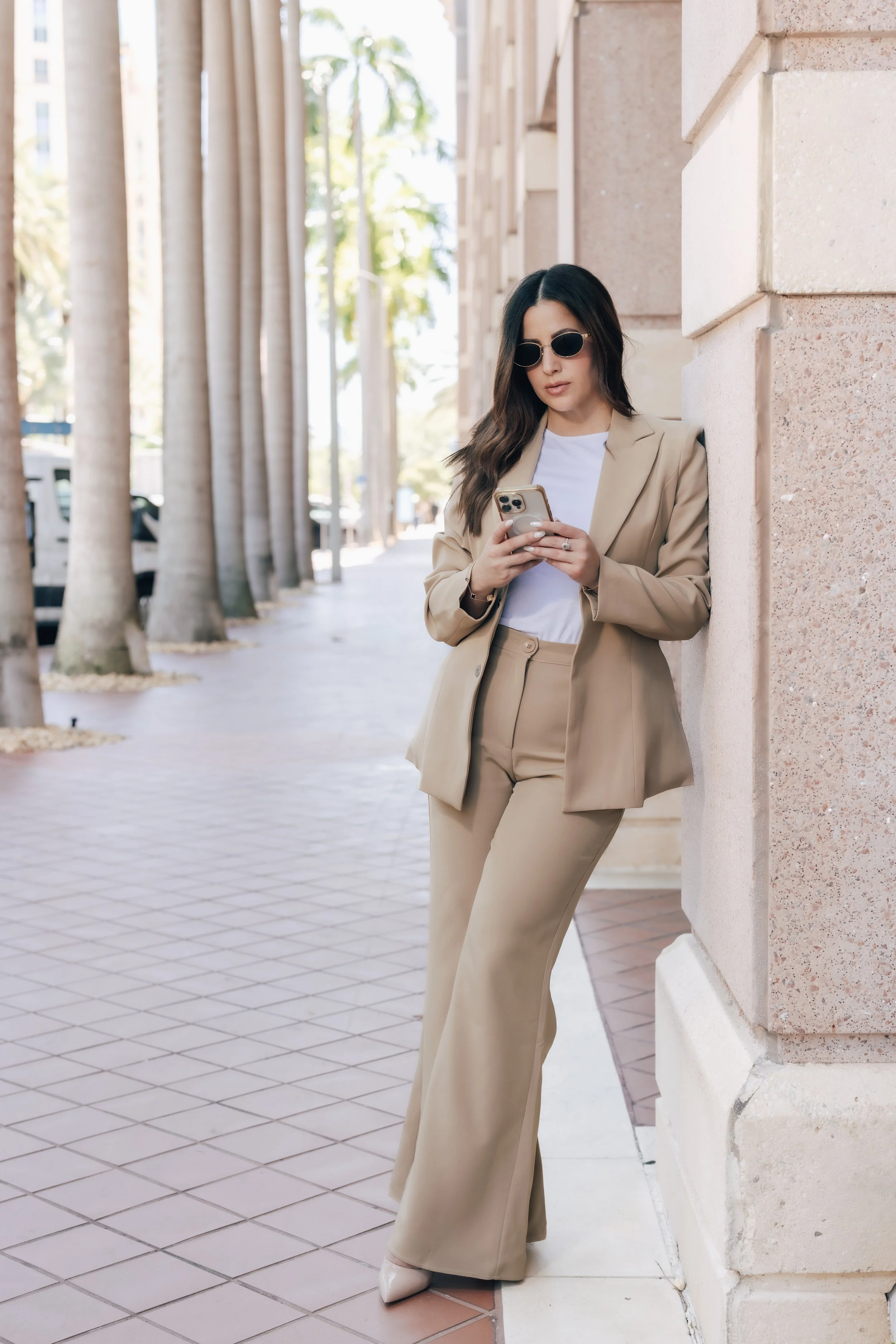 A woman in a tan suit with sunglasses leaning against a beige stone building on a sidewalk, looking at her phone.