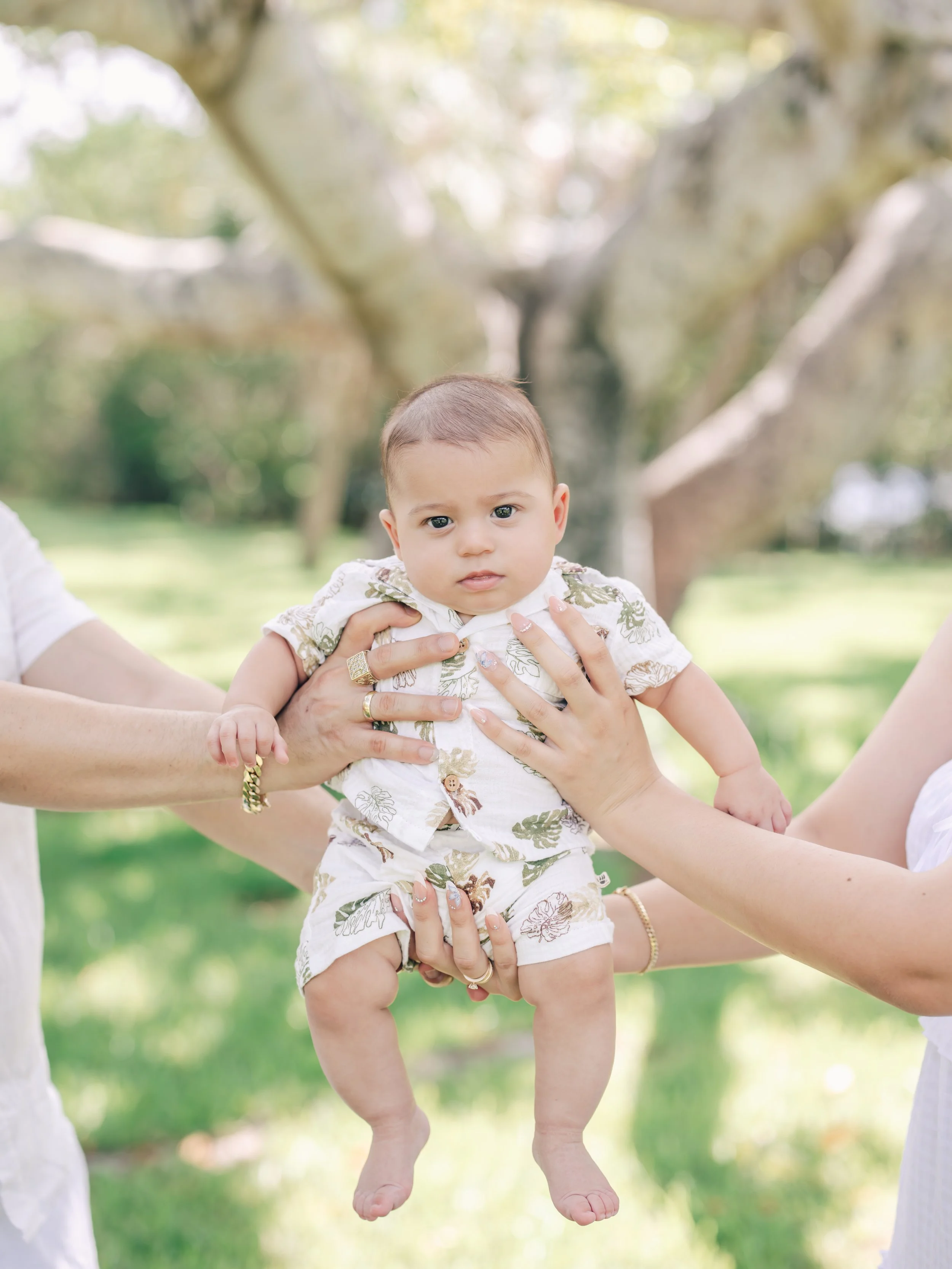 A baby in a floral outfit being held up outdoors with greenery and trees in the background.