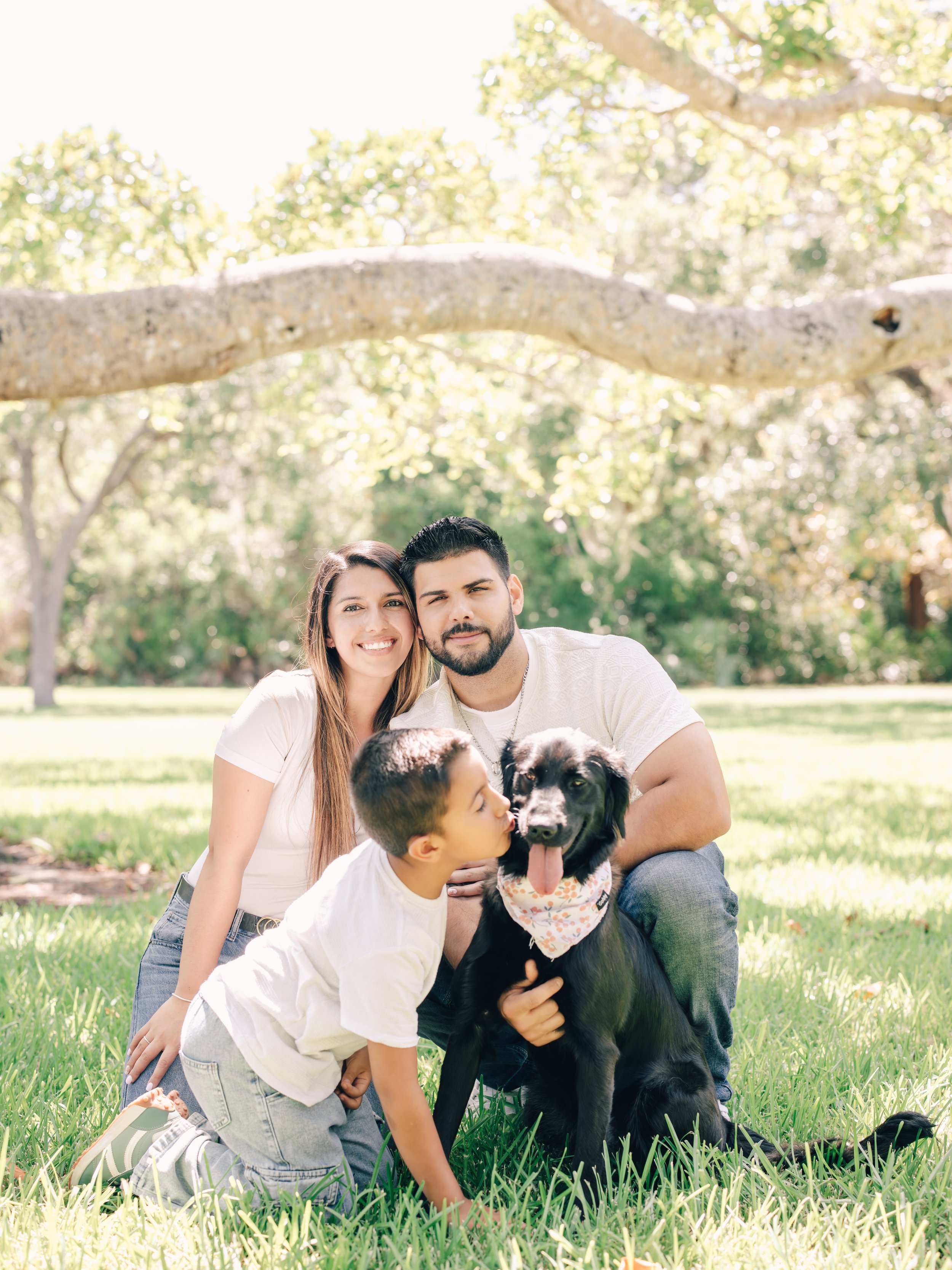 Young couple with a dog and a young boy in a park with green grass and trees, smiling and enjoying a sunny day.