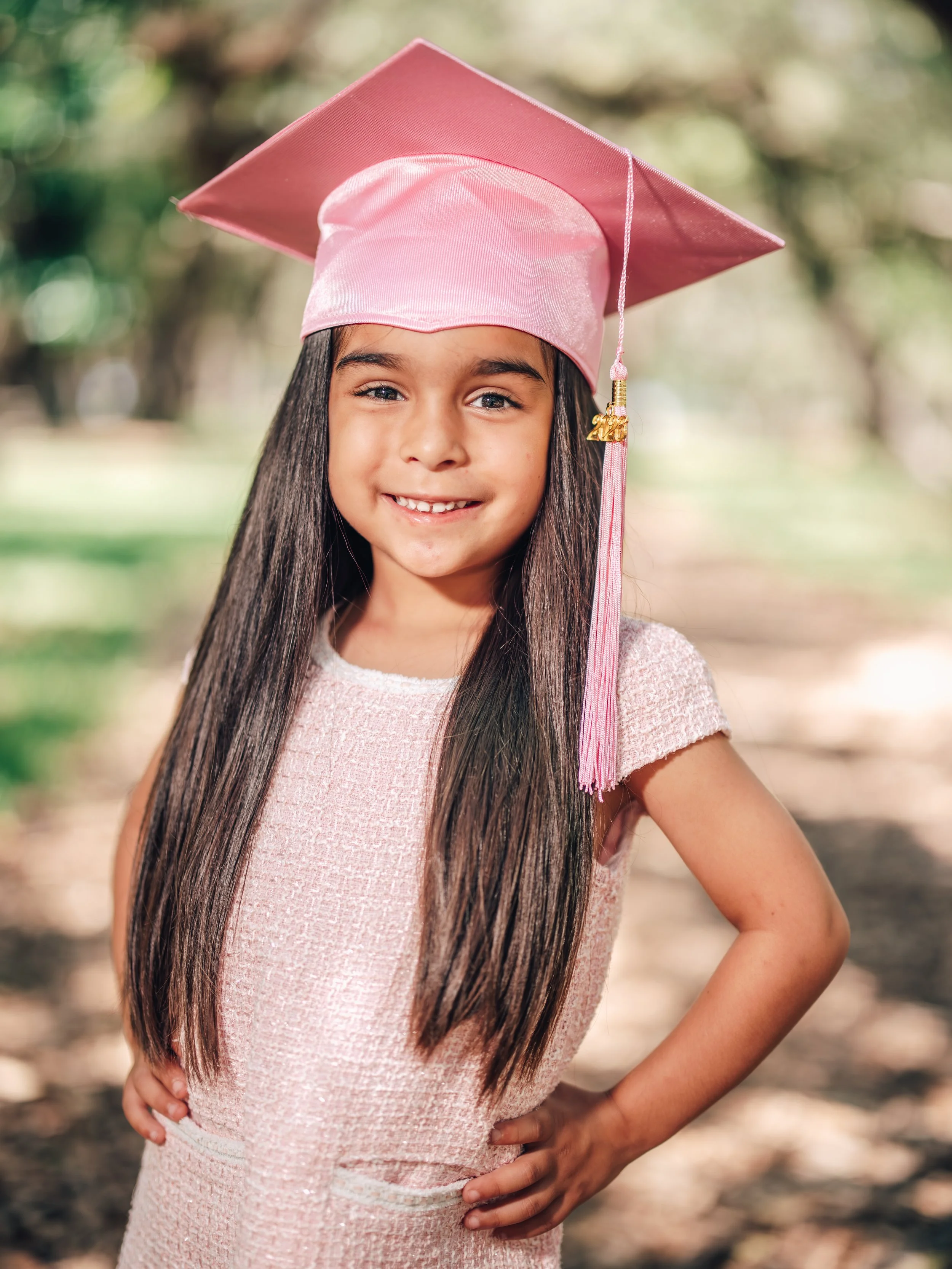 A young girl with long dark hair wearing a pink graduation cap and dress, smiling outdoors with a blurred natural background.