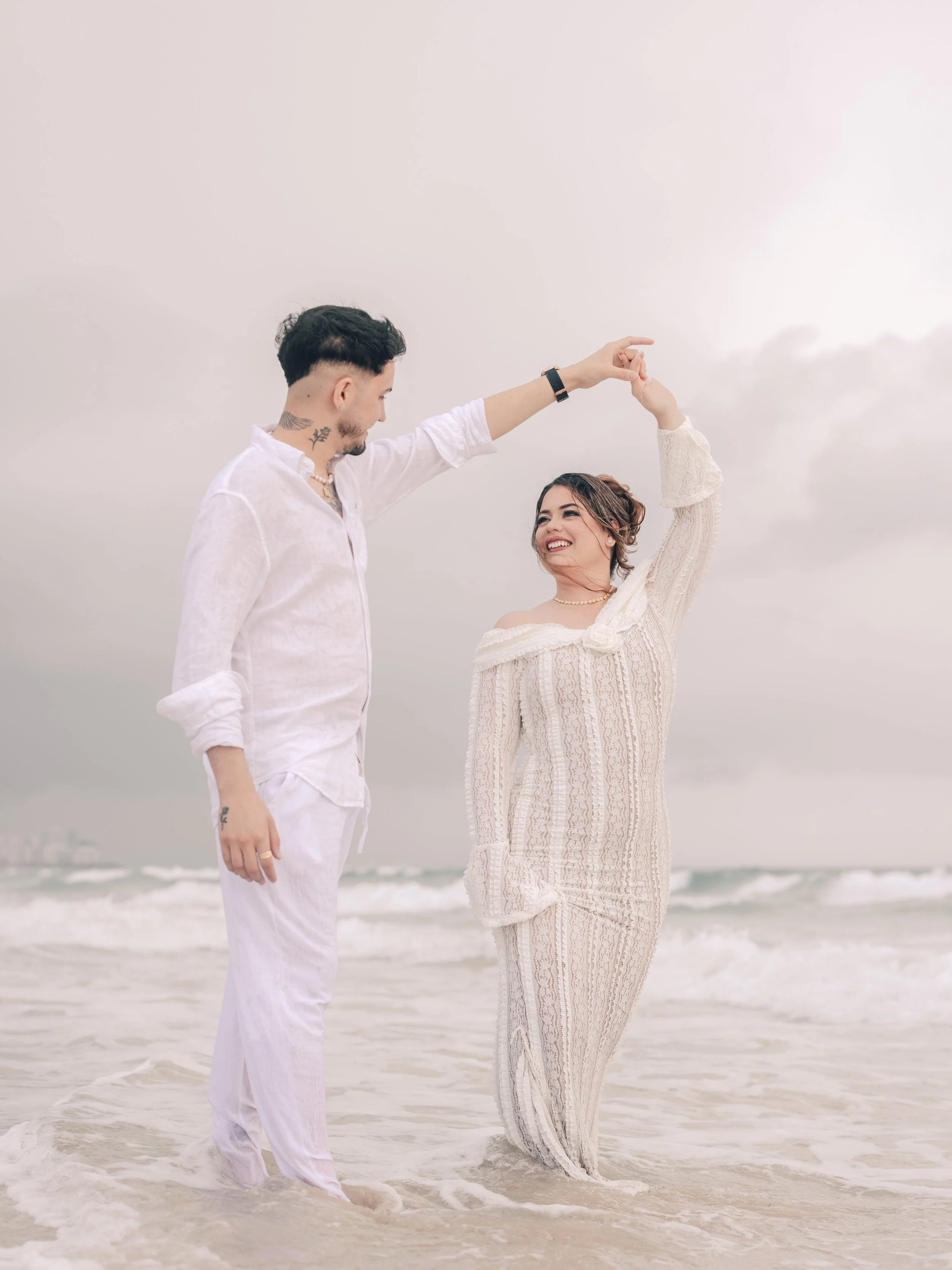 A couple dancing on a beach with ocean waves in the background, the man wearing a white shirt and pants, and the woman in a light-colored dress, both smiling and holding hands.