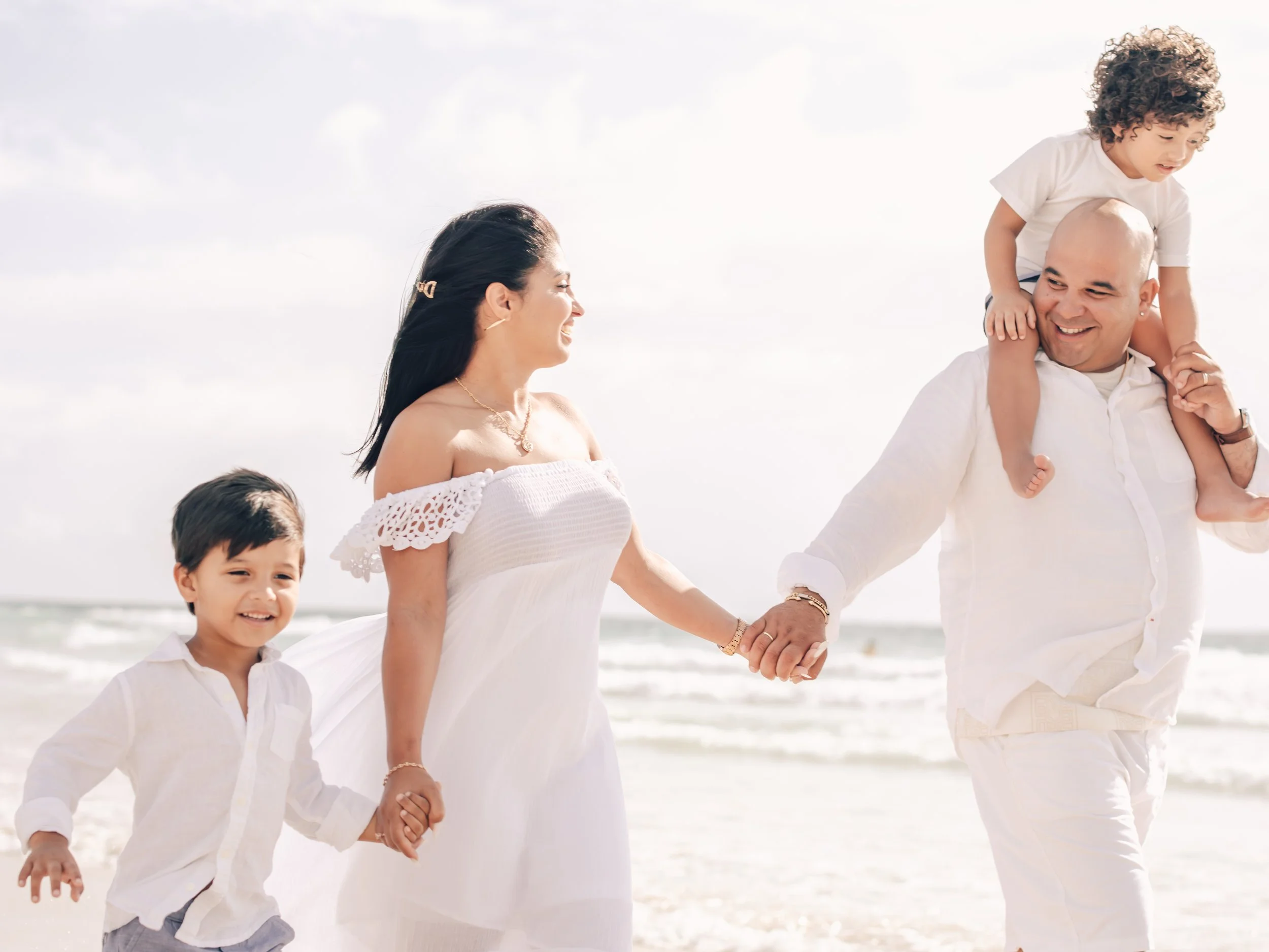 A family walking on the beach, holding hands, with the ocean and sky in the background. The woman is smiling and wearing a white off-the-shoulder dress, the man is carrying a young child on his shoulders, and two other children are walking alongside.