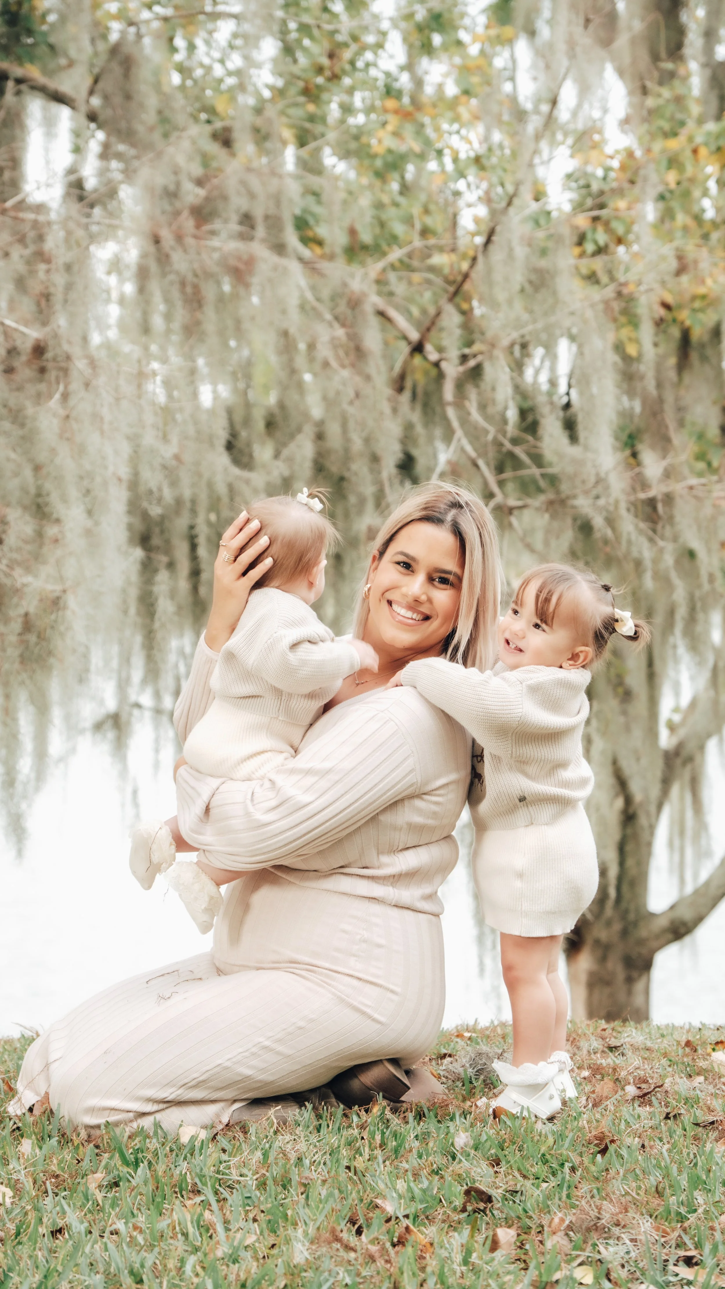 A woman kneels on a grassy area hugging two young girls near a tree with hanging moss, by a body of water, in an outdoor setting during autumn.
