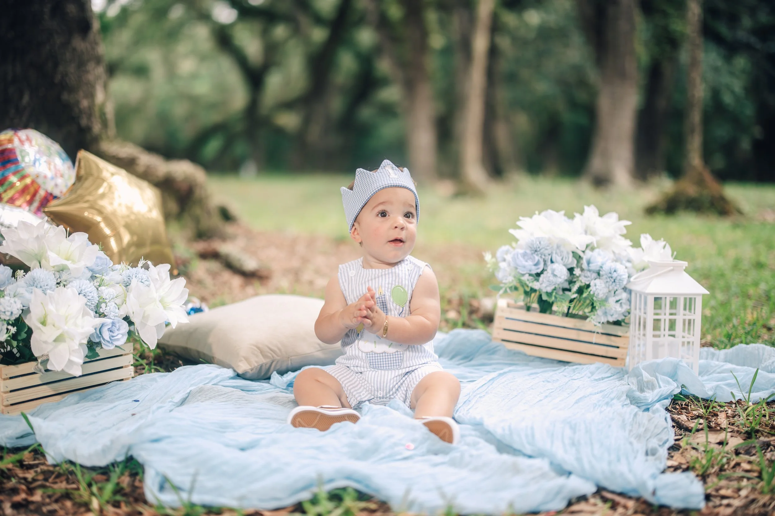 Baby girl sitting on a light blue blanket outdoors surrounded by white and blue flowers and balloons, with trees in the background.