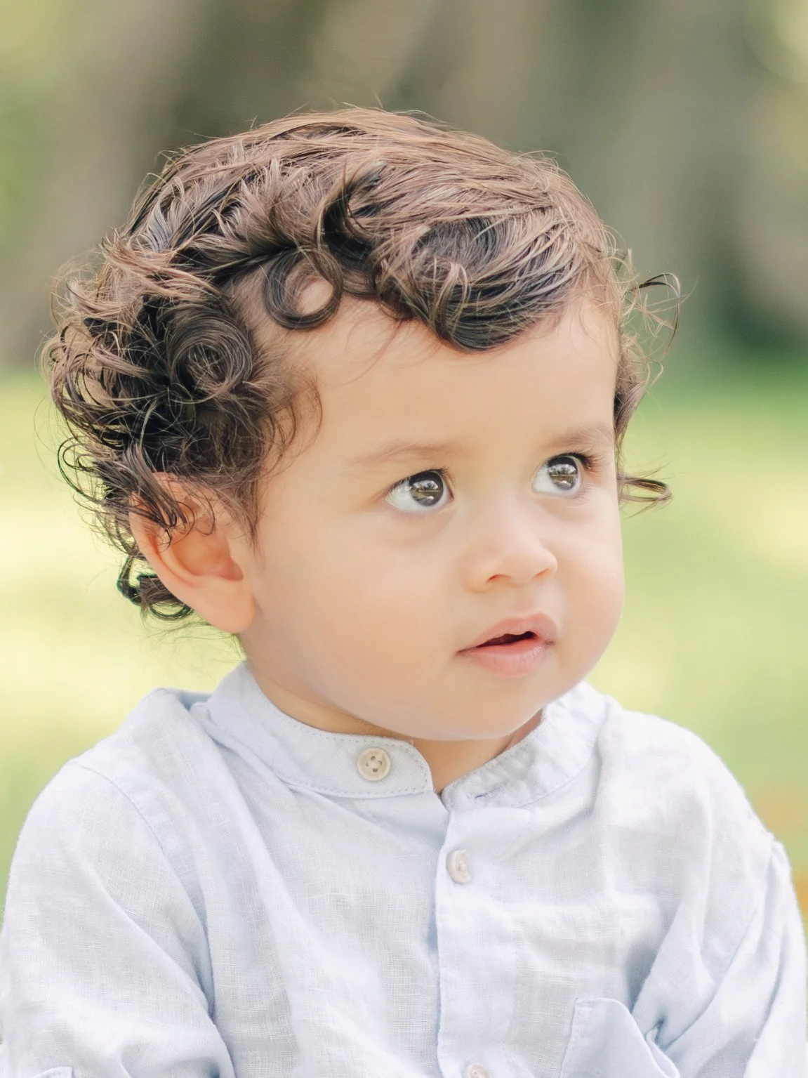 Close-up of a young child with curly brown hair and brown eyes, wearing a white shirt, outdoors with blurry green background.