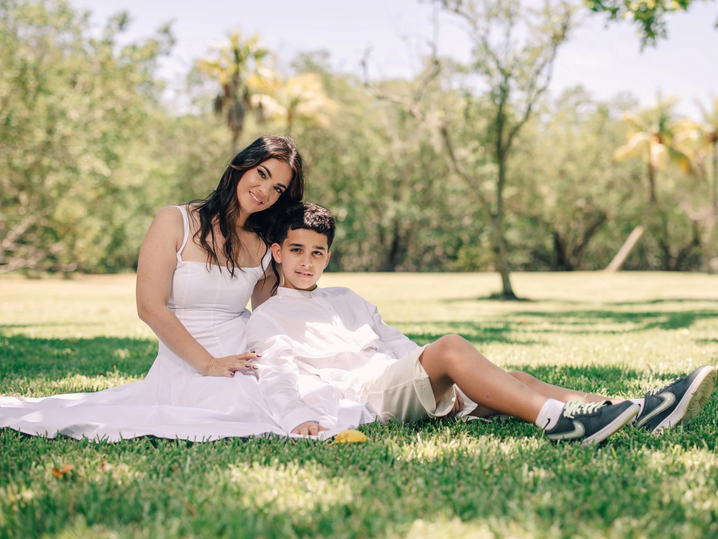 A woman with dark hair and a young boy with short dark hair sitting outdoors on a grassy area with trees in the background, enjoying a sunny day.