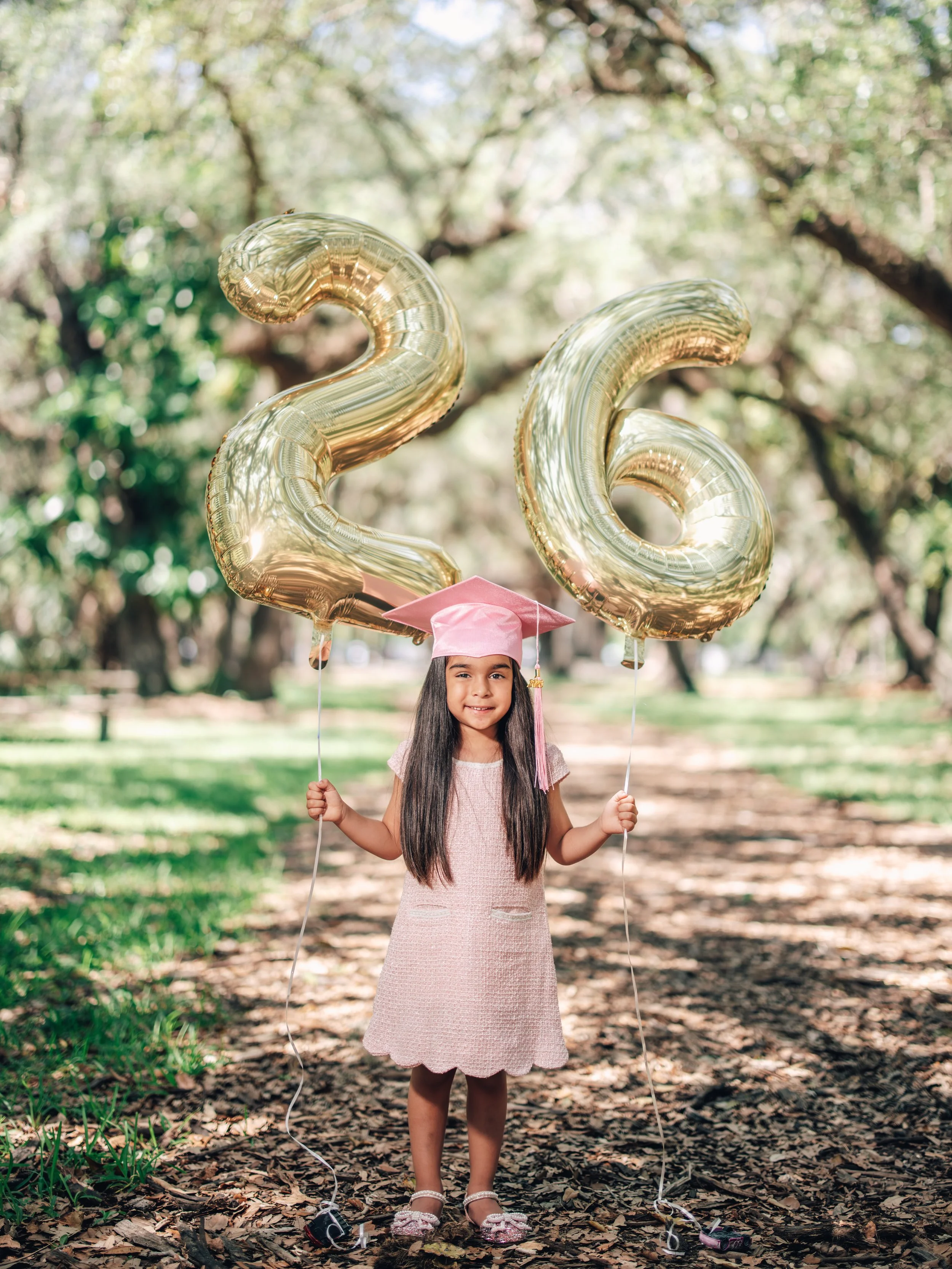 Young girl in a pink dress and matching graduation cap holding two large gold Christmas-shaped balloons in a park with trees and fallen leaves.