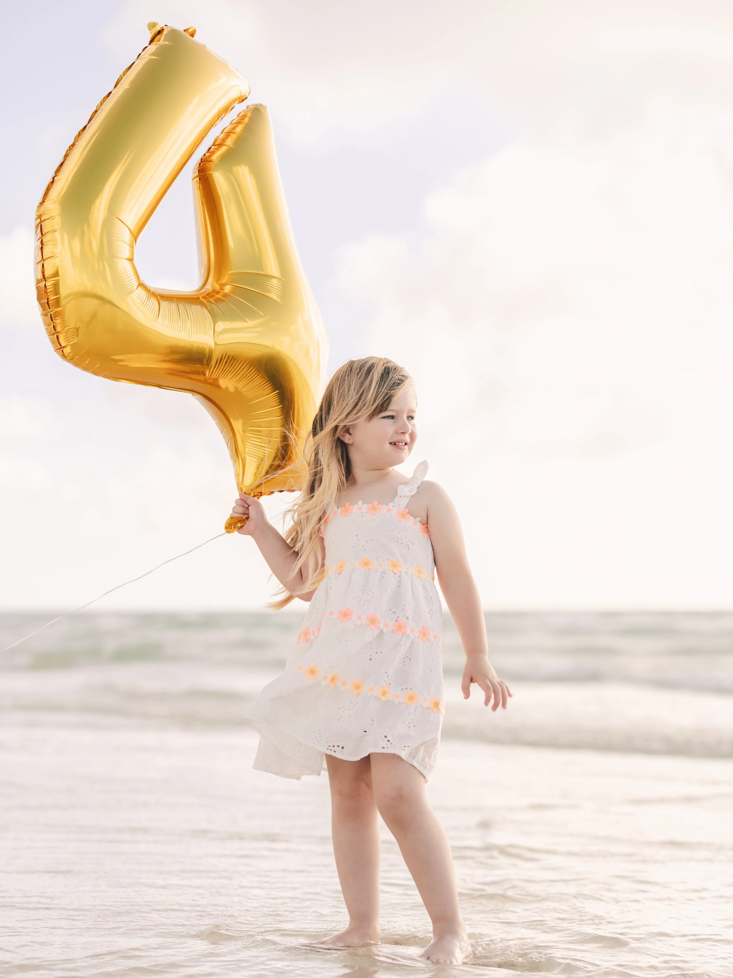 A young girl standing in shallow ocean water holding a large gold balloon shaped like the number 4. She is wearing a white dress with pink and yellow flower details and looks out towards the distance.