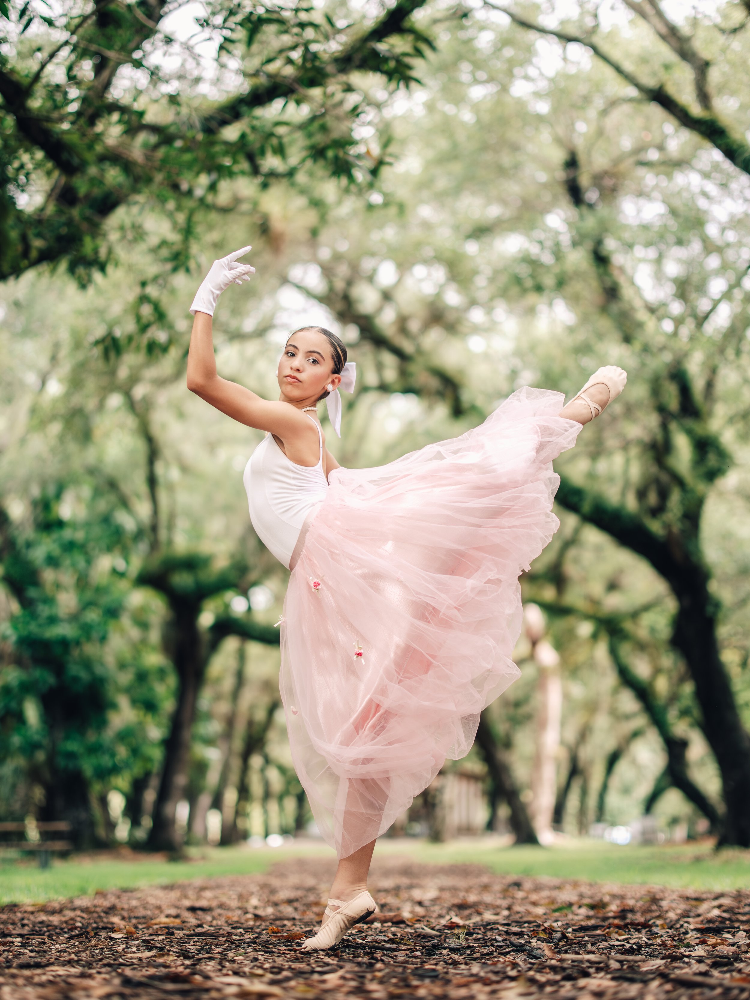 A ballerina in a pink tutu performing a dance pose on a wooded park path covered with fallen leaves, with trees and green foliage in the background.
