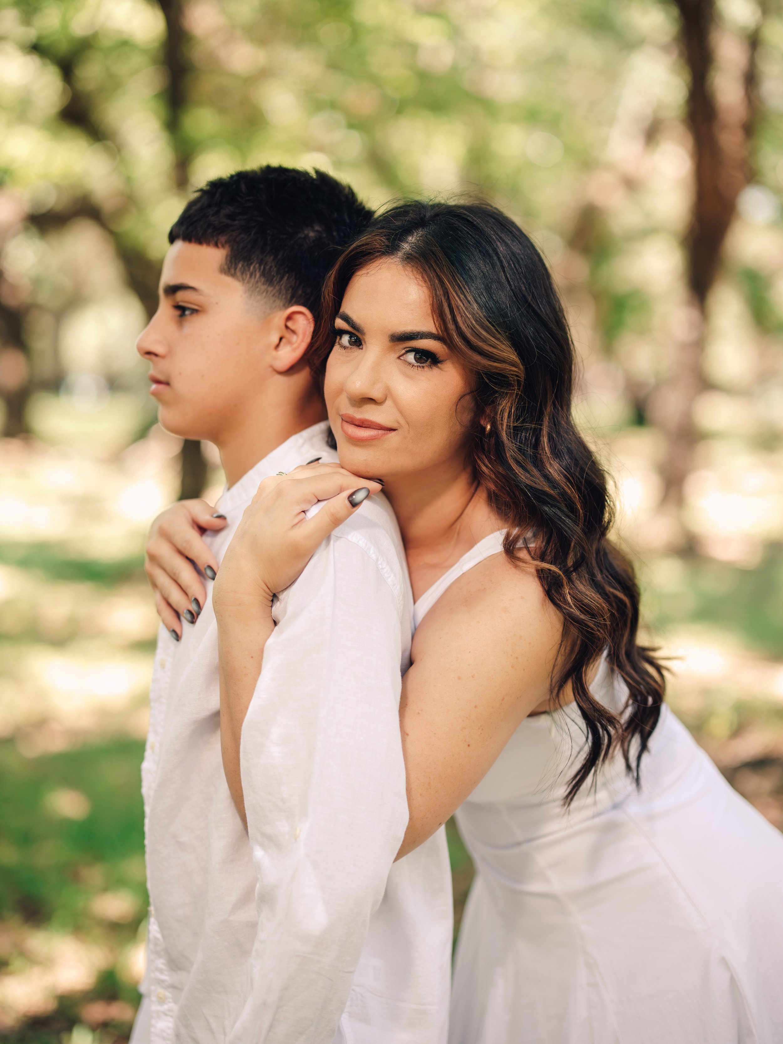 A woman with long dark hair and makeup posing with a young man outdoors during daytime, both wearing white shirts, with sunlight filtering through green trees in the background.