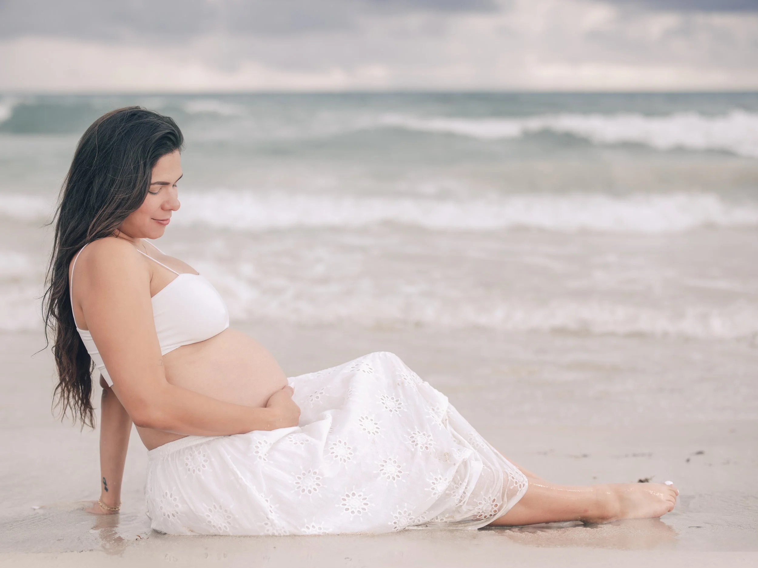 A pregnant woman with long dark hair sitting on a beach, wearing a white dress, with ocean waves in the background.