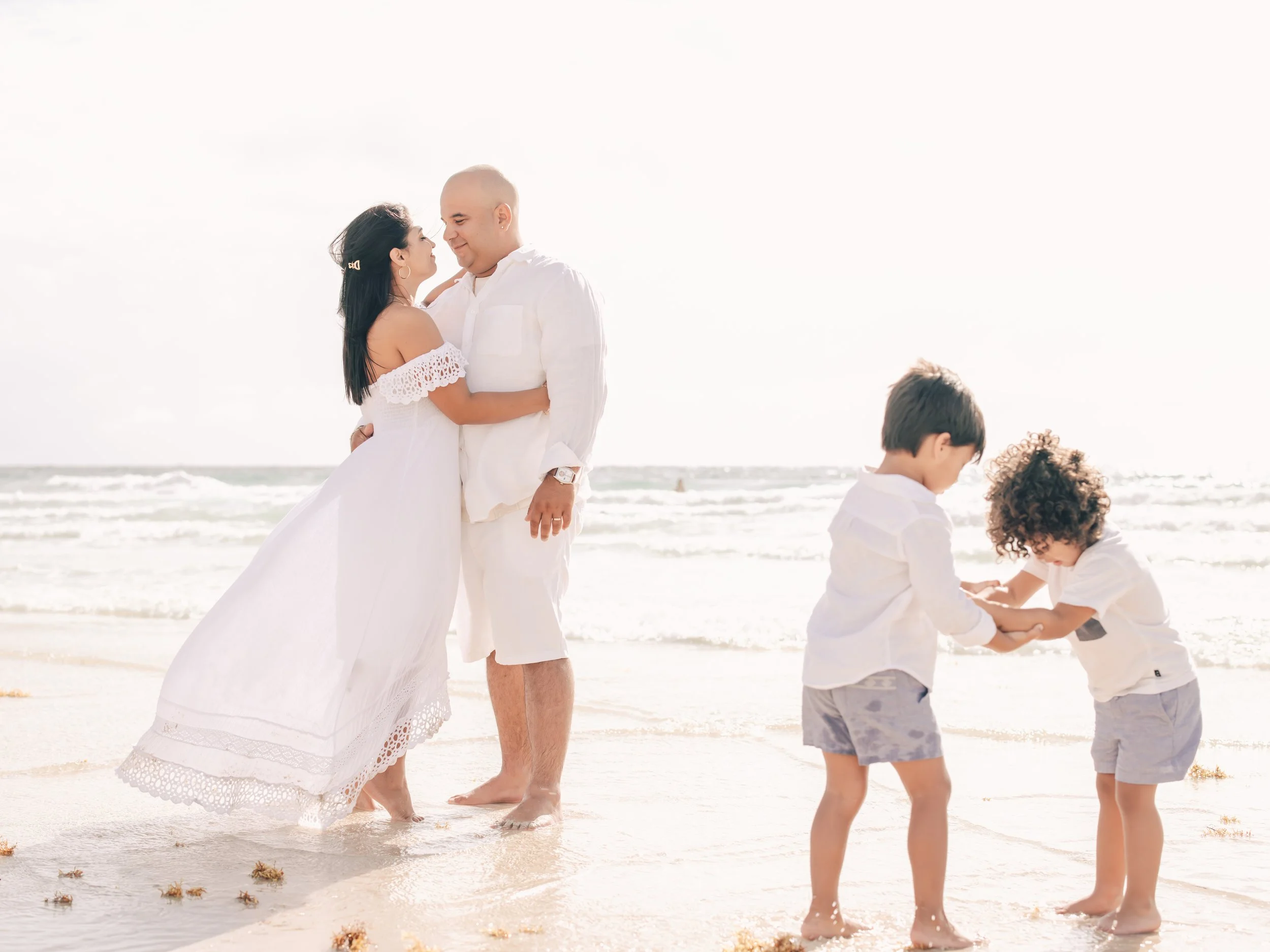 A couple dressed in white standing and smiling at each other on a beach, with children playing in the sand nearby.