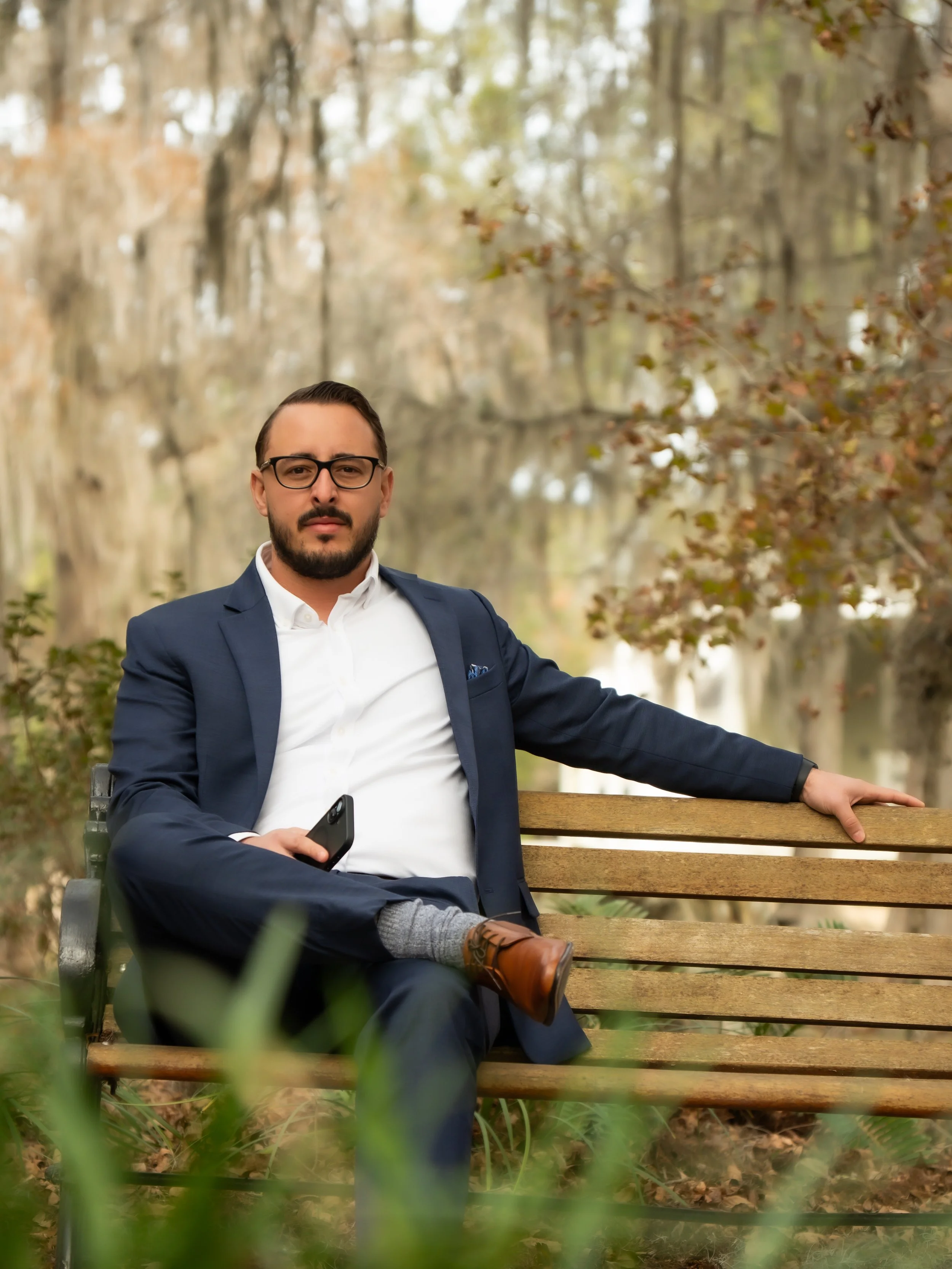 A man in a business suit sitting on a park bench, holding a smartphone, with trees and foliage in the background at Silver Springs Park in florida