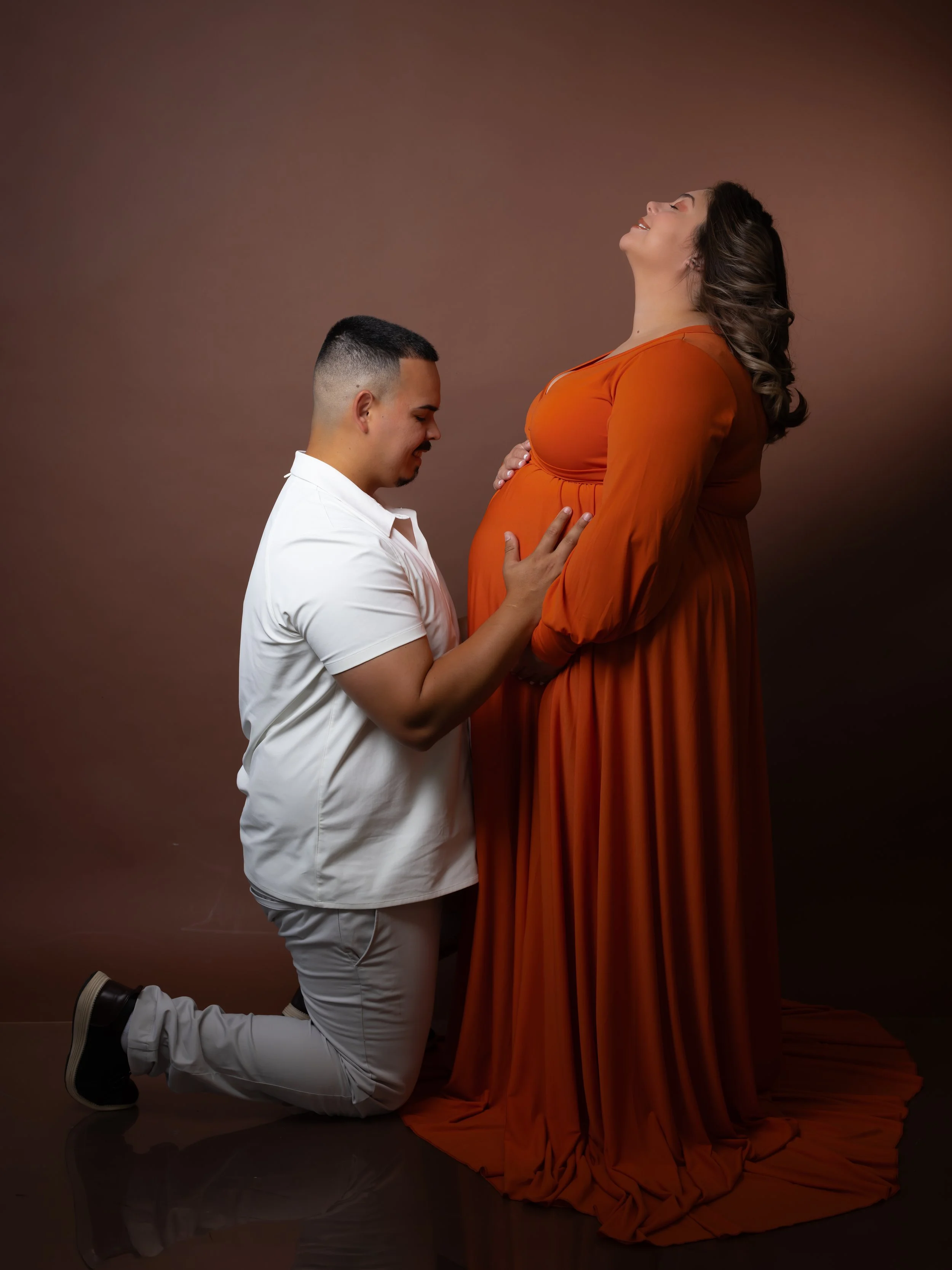 A man in white kneeling with hands on pregnant woman's belly, holding her hand, and she is wearing an orange dress with her head tilted back.
