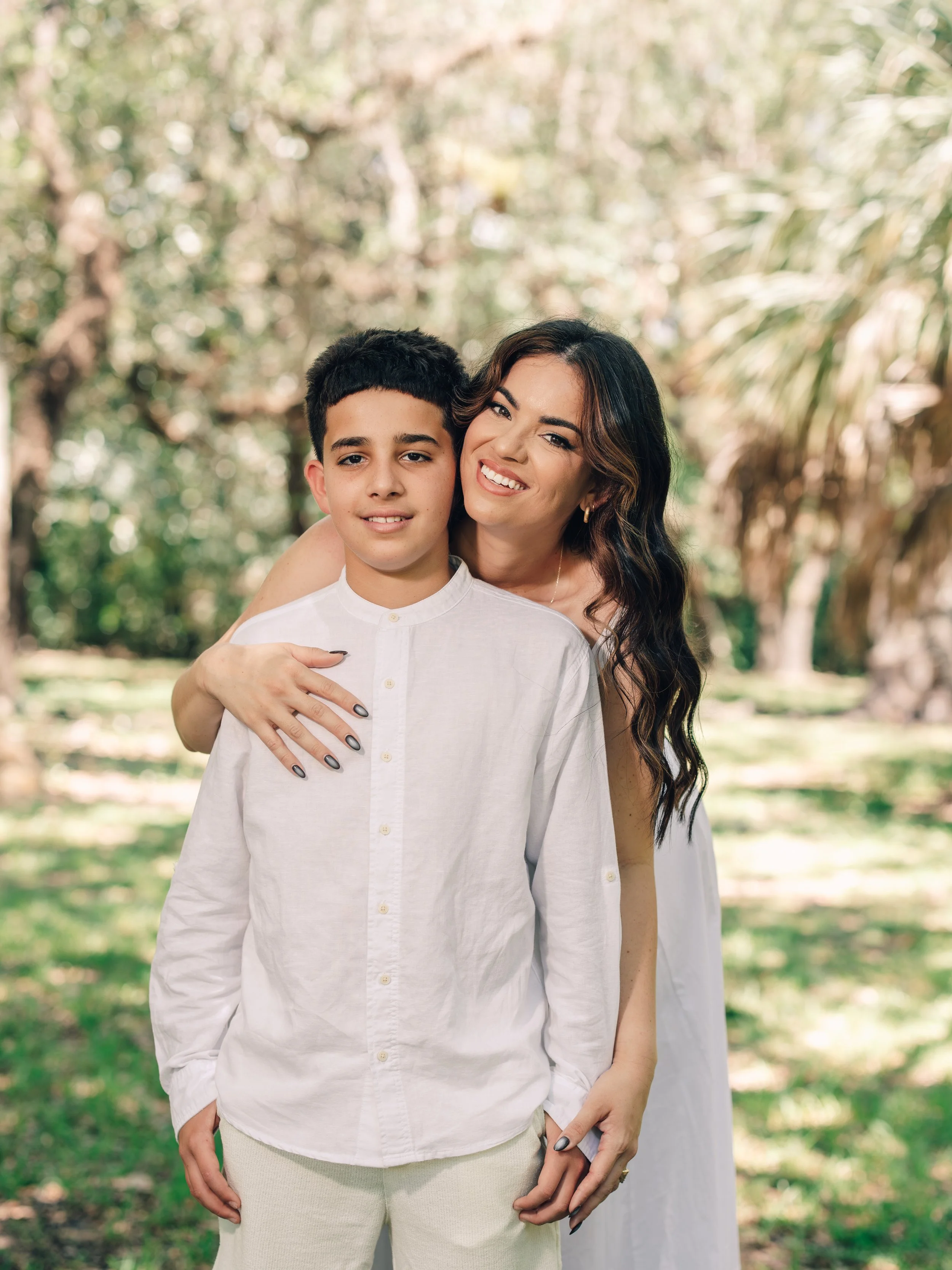 A woman with long, dark, wavy hair and a boy with short, dark hair standing outdoors in a green, wooded area. The woman is smiling and winking, with her arm around the boy, who is looking at the camera with a neutral expression.
