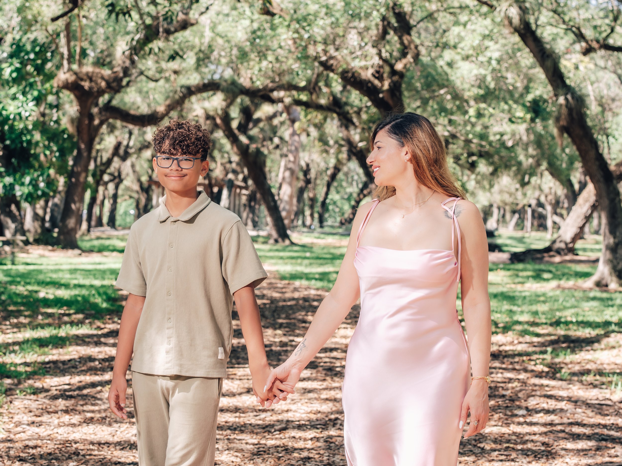 A woman with long, brown hair and a pink satin dress holding hands with a young man with curly brown hair, glasses, and a beige shirt, walking outdoors in a park with large trees and green grass.