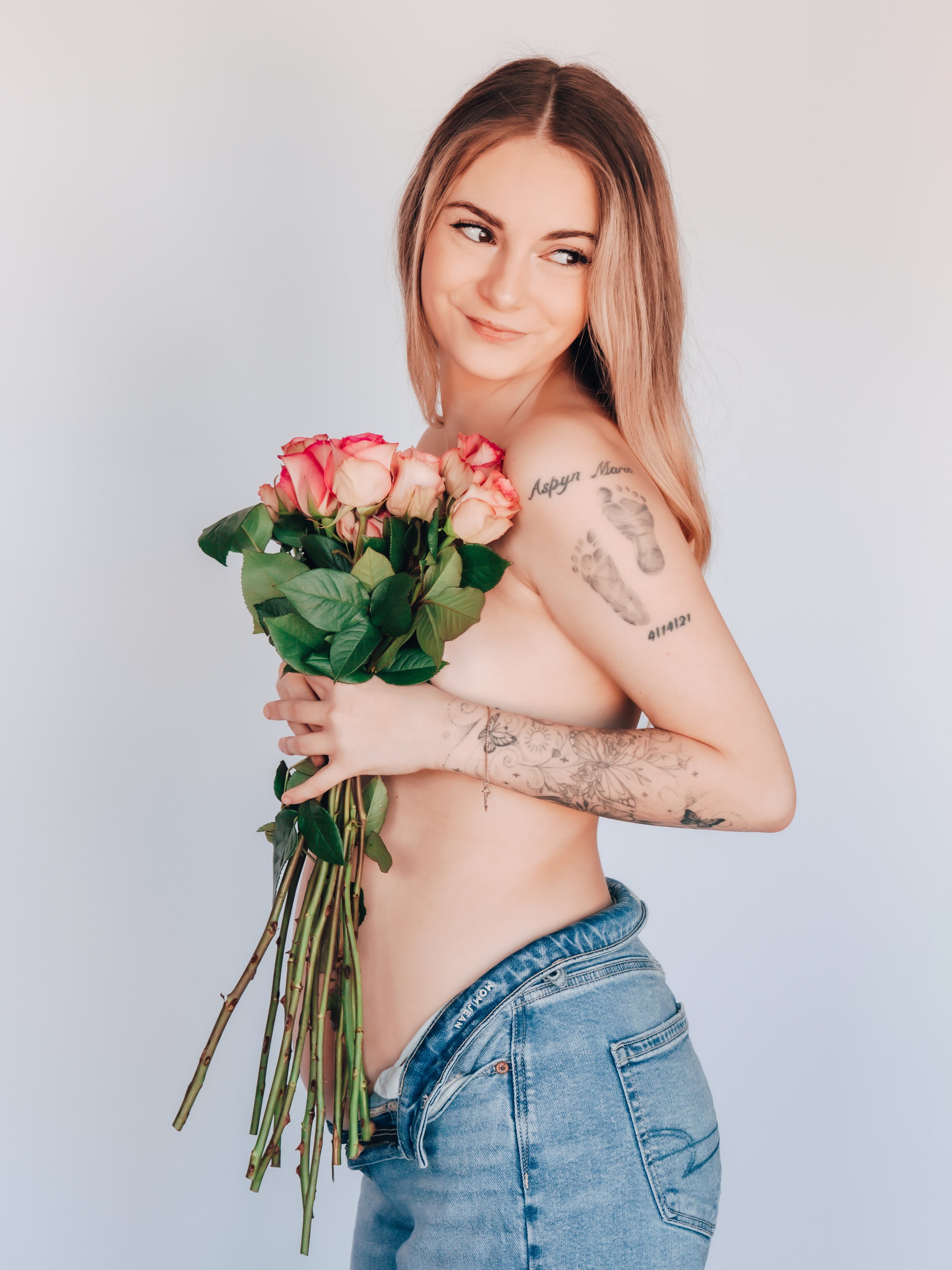 A young woman with long, light brown hair holding a bouquet of pink roses, topless with tattoos on her left arm and shoulder, wearing blue jeans, and smiling with a playful expression.