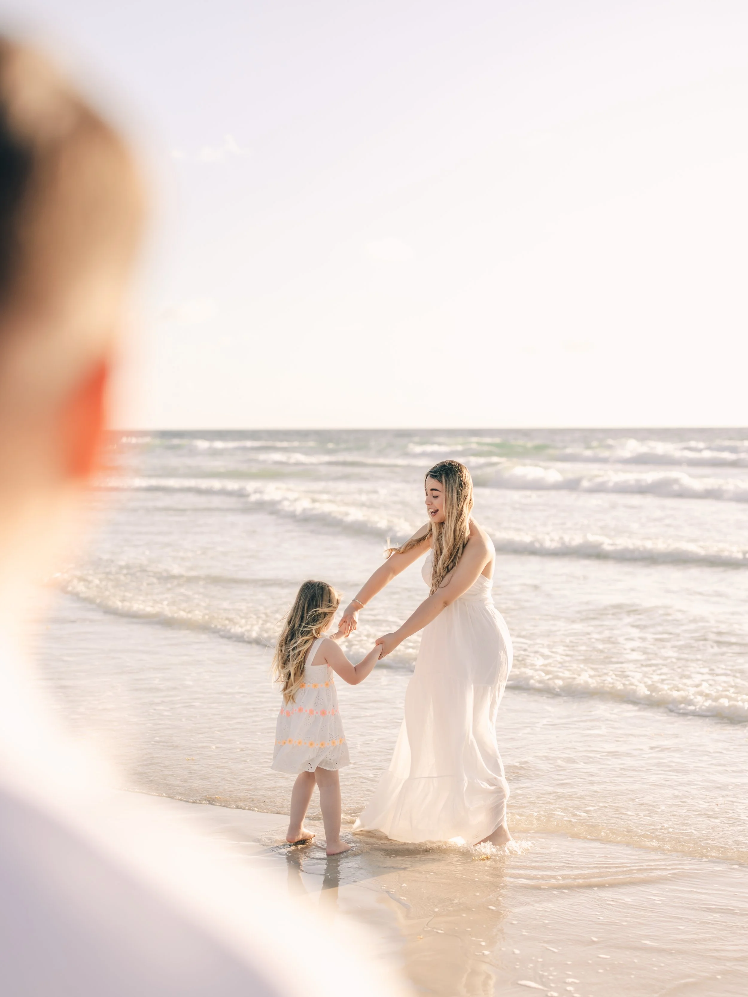 A woman in a white dress and a young girl in a white dress with orange details holding hands and standing in shallow ocean water at the beach.
