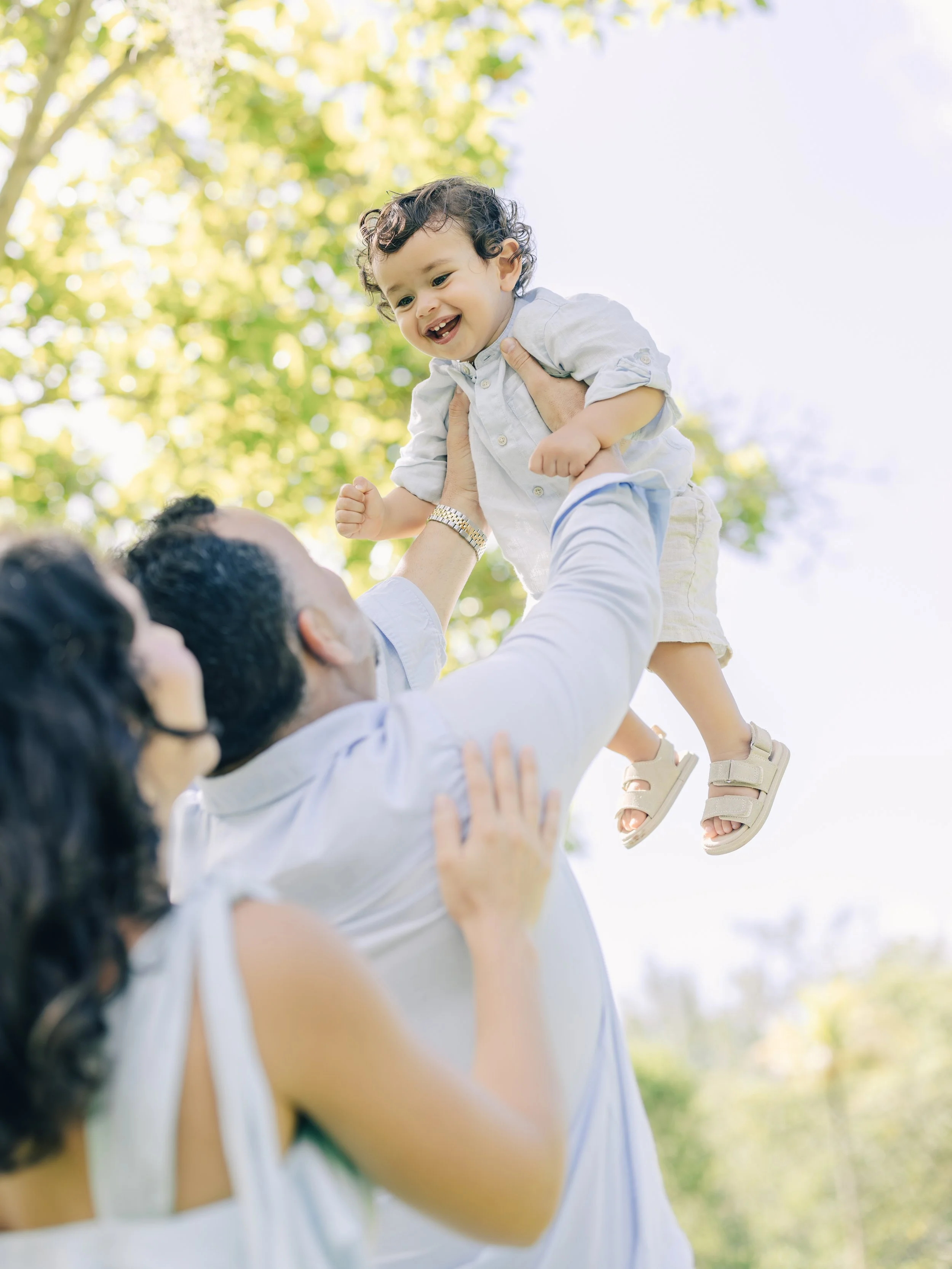 Child being held up in a park by an adult, laughing and looking happy with trees and sunlight in the background.