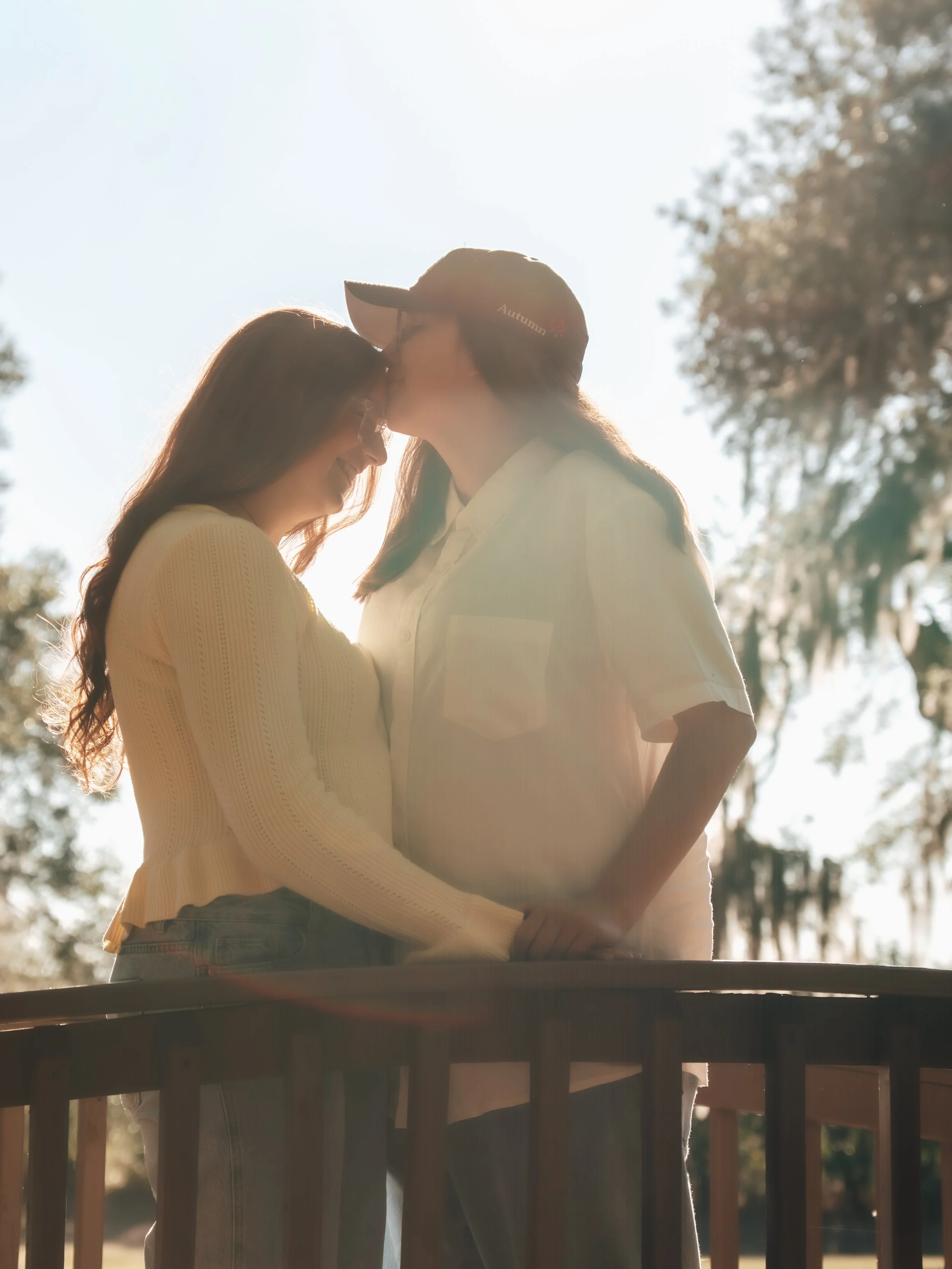 Two women sharing an intimate moment, one kissing the other's forehead, backlit by sunlight outdoors at a park in central florida.