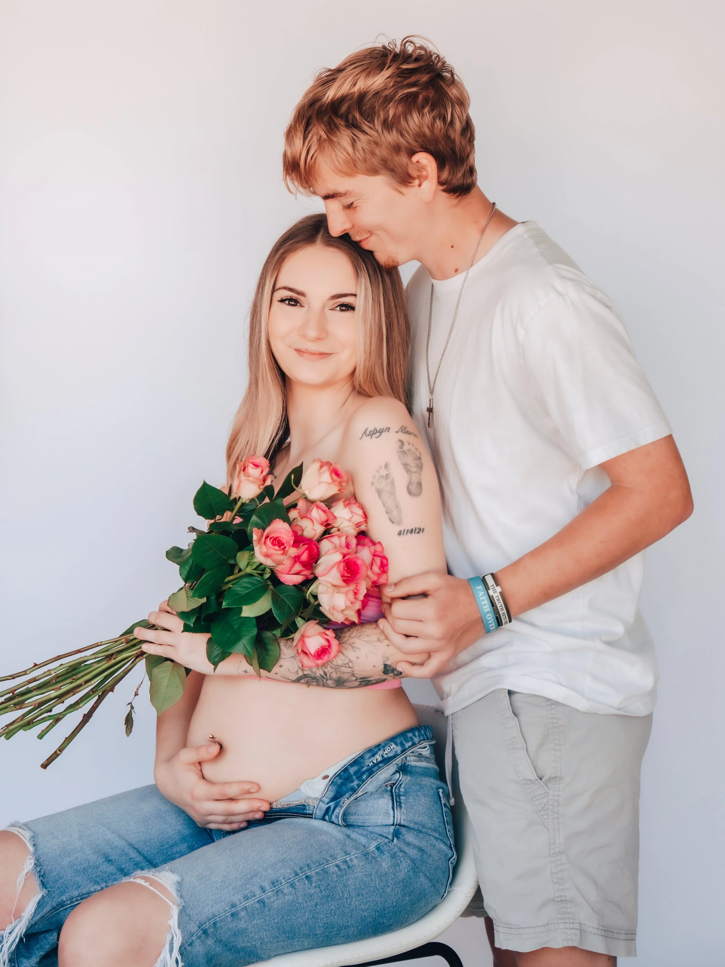 A pregnant woman with tattoos sitting on a chair holding a bouquet of pink roses, smiling. A man standing close to her, gently holding her arm, smiling, with his head resting on her forehead.