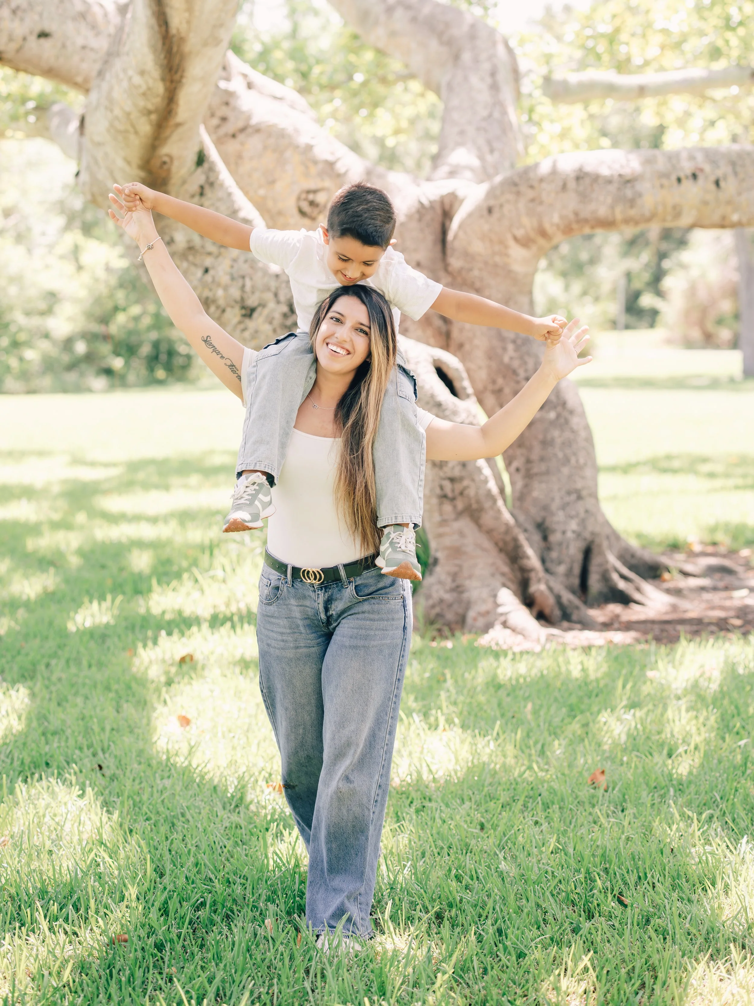 A woman carrying a young boy on her shoulders in a park with green grass and trees.