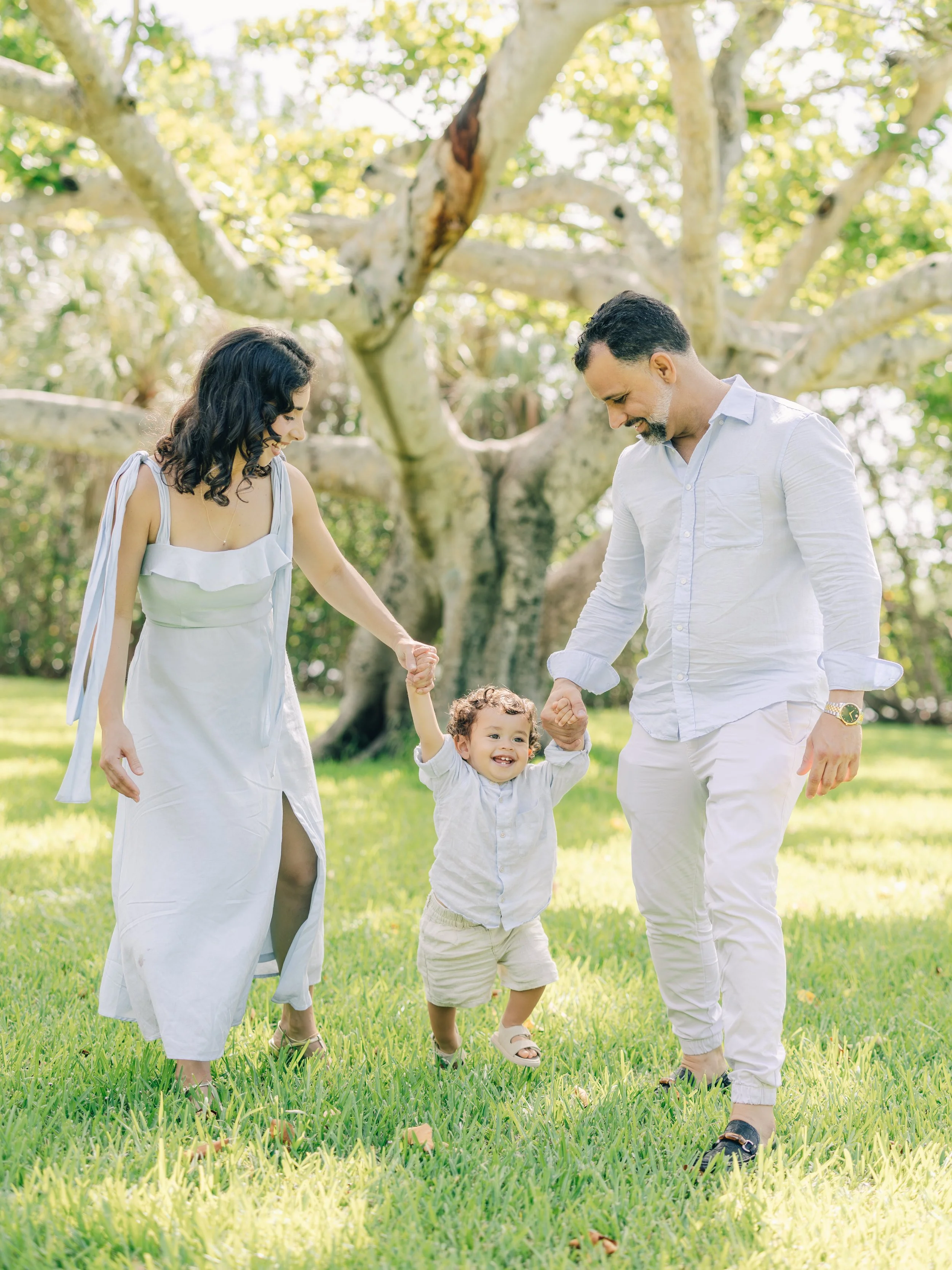 A family of three holding hands and walking on a grassy field with a large tree in the background. The mother, father, and young child are smiling and enjoying a sunny day outdoors.