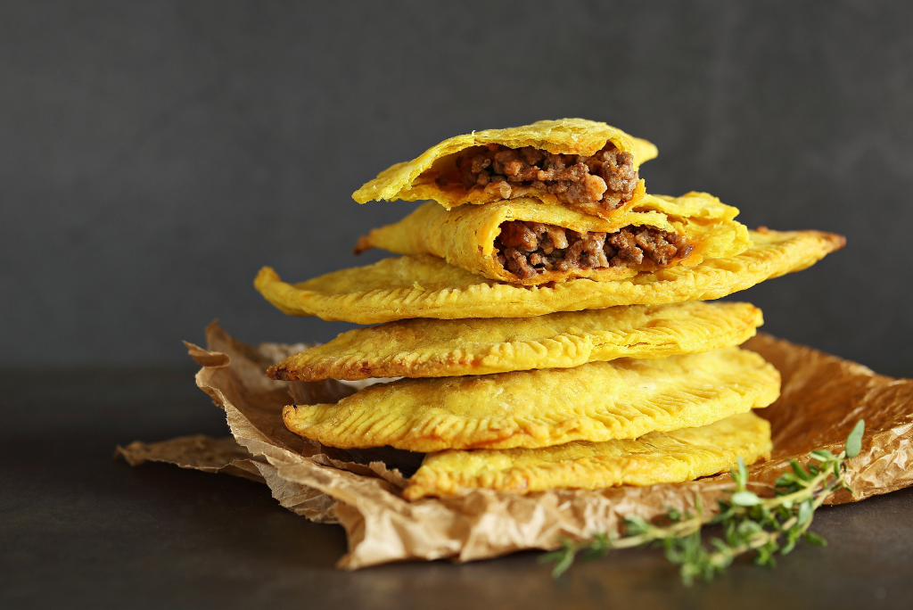 Stack of five fried meat pies with one cut open to reveal beef filling, on crumpled parchment paper with sprigs of thyme.