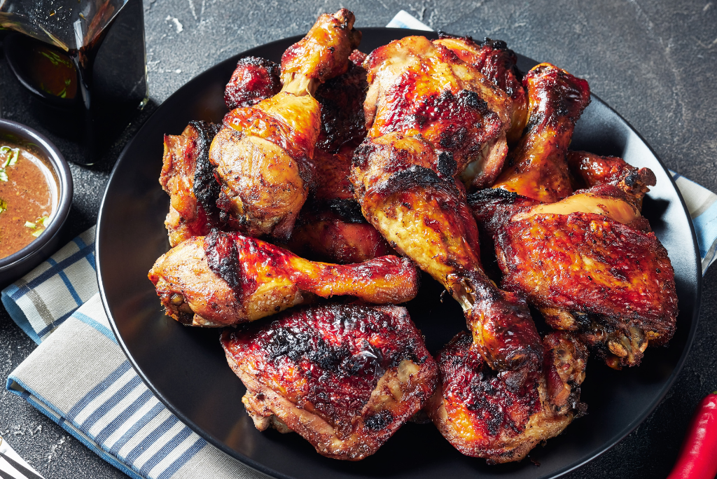 A black plate filled with grilled chicken drumsticks and thighs, some with a charred appearance, served on a dark surface with a salt and pepper shaker, a small bowl of dipping sauce, and a glass of soda nearby.