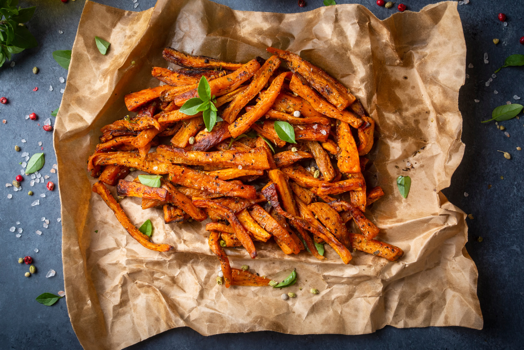 A serving of roasted sweet potato fries garnished with fresh basil leaves, served on brown parchment paper with scattered peppercorns and seasoning on a dark surface.