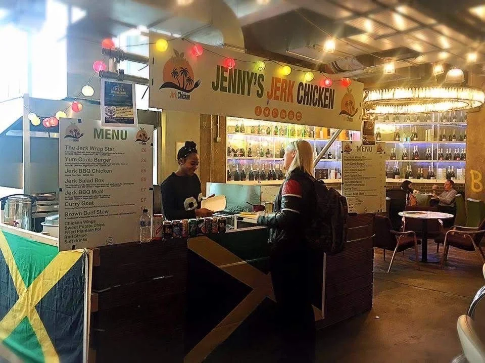 Jenna's Jerk Chicken food stall inside a restaurant, with a customer ordering, menu signs, and Jamaican flag in the foreground.